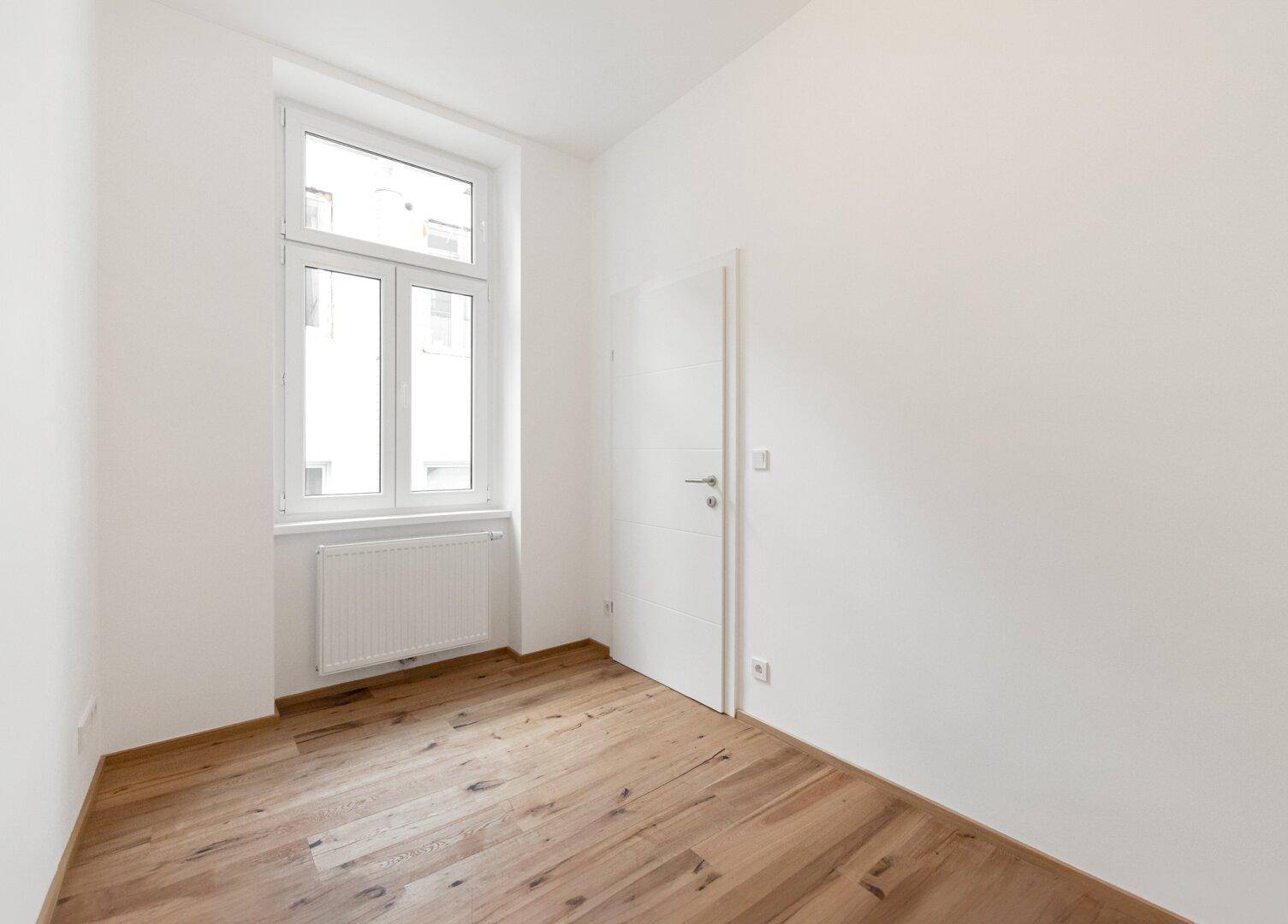 Leeres Schlafzimmer mit Holzboden und einem großen Fenster, das viel Tageslicht bietet.