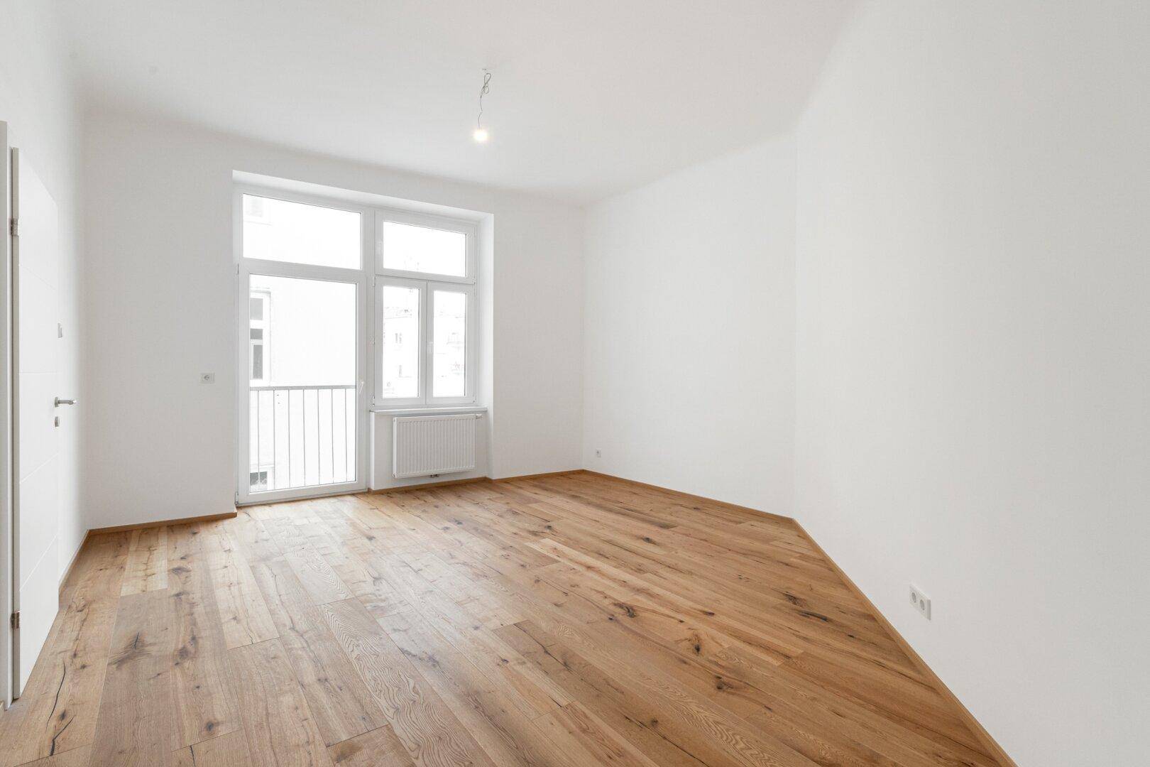 Geräumiges, leeres Wohnzimmer mit Holzboden und großen Fenstern, die viel Tageslicht hereinlassen.