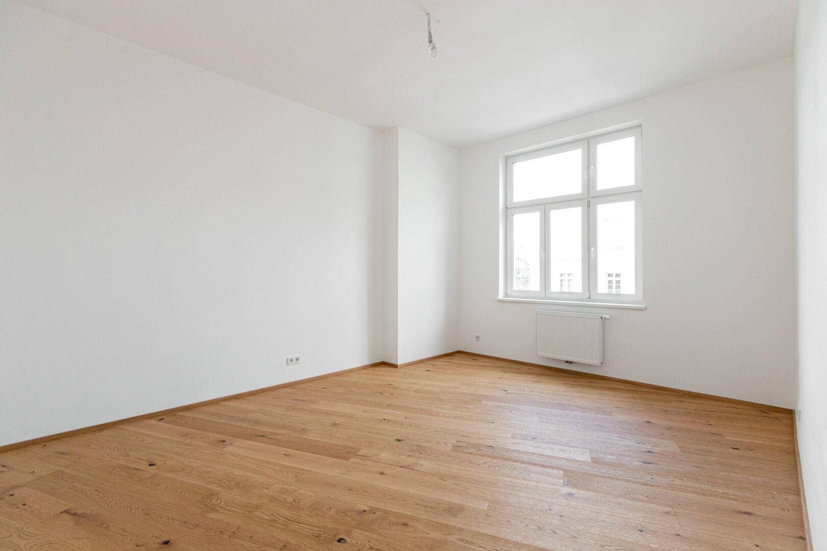 Leeres Zimmer mit Parkettboden, weißen Wänden und einem großen Fenster, ideal als Schlafzimmer.