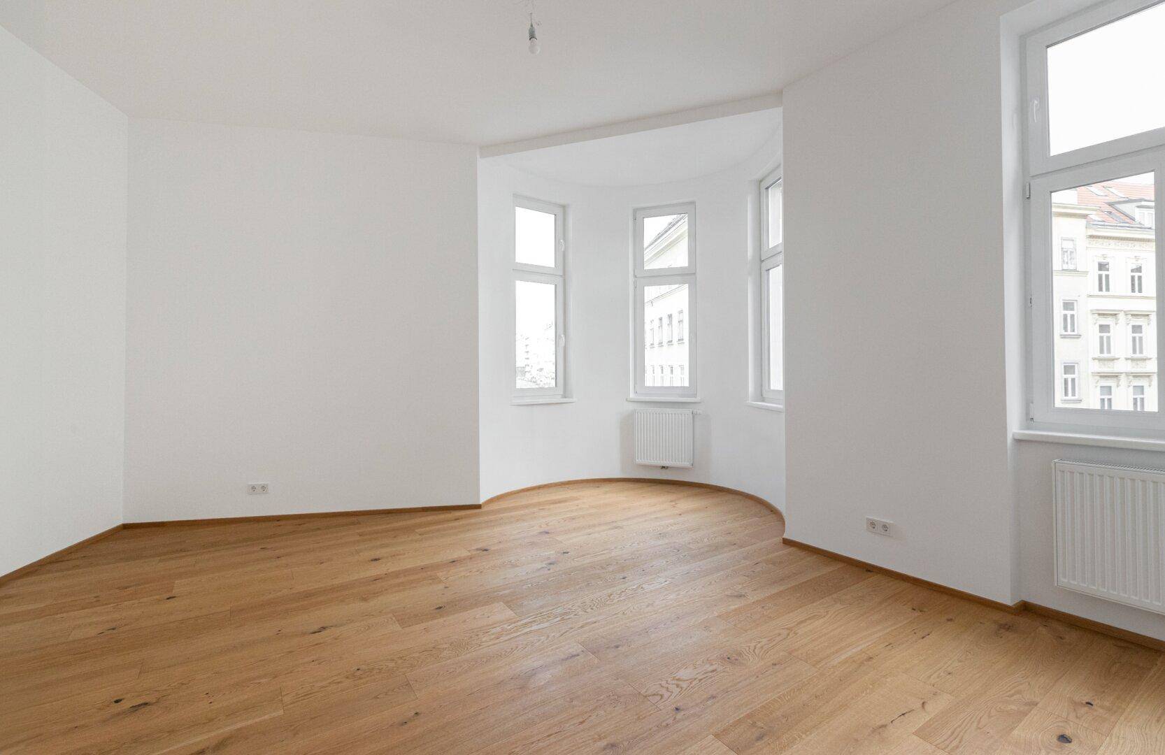 Geräumiges, leeres Zimmer mit Parkettboden, weißen Wänden und einem Erkerfenster.
