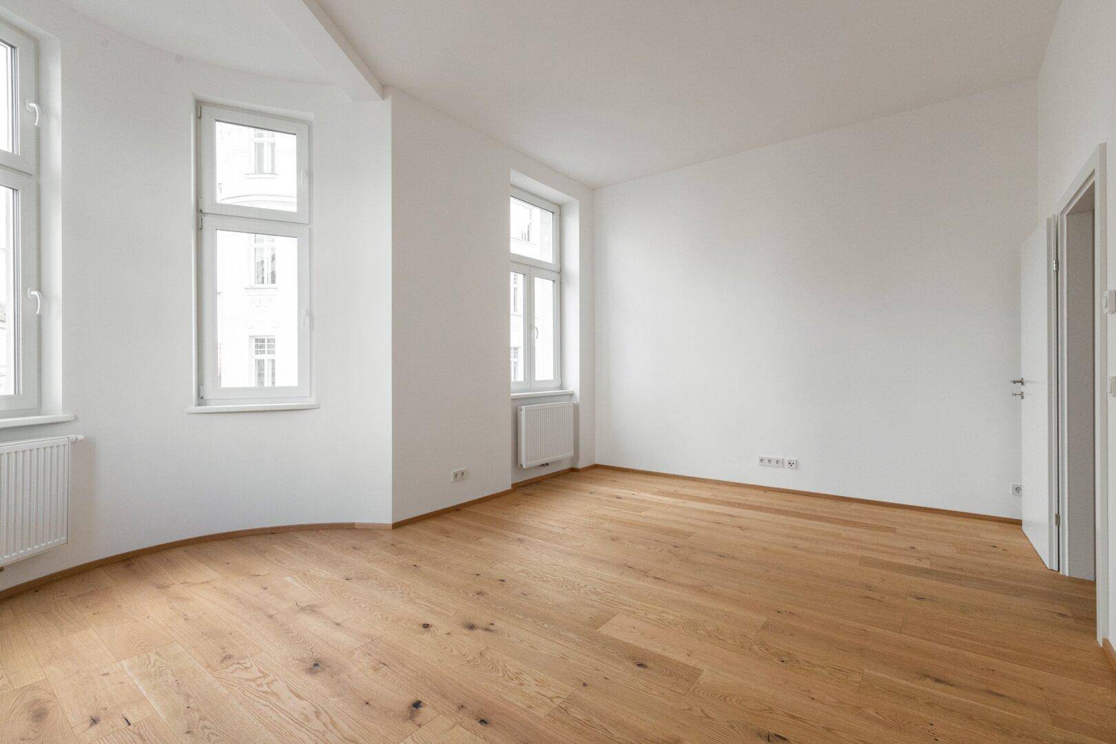 Helles, leeres Zimmer mit Parkettboden, weißen Wänden und hohen Fenstern.