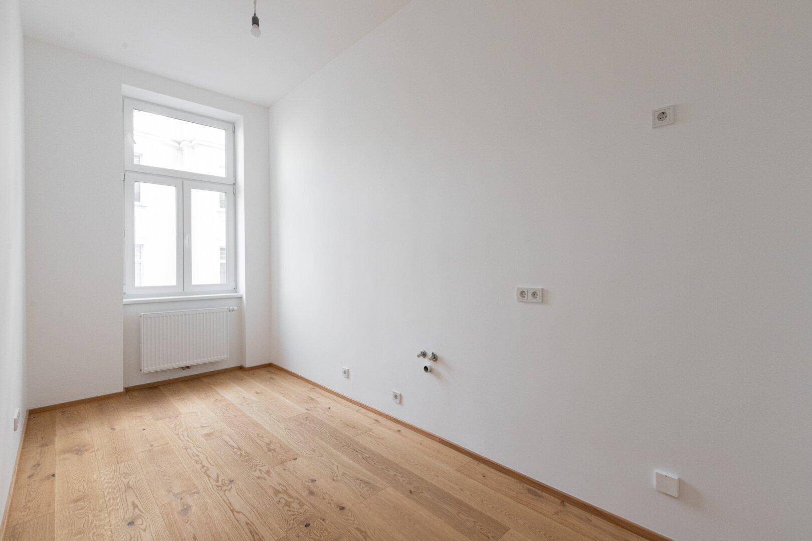 Leerer Küchenraum mit Parkettboden, weißen Wänden und Anschlüssen für eine Einbauküche.