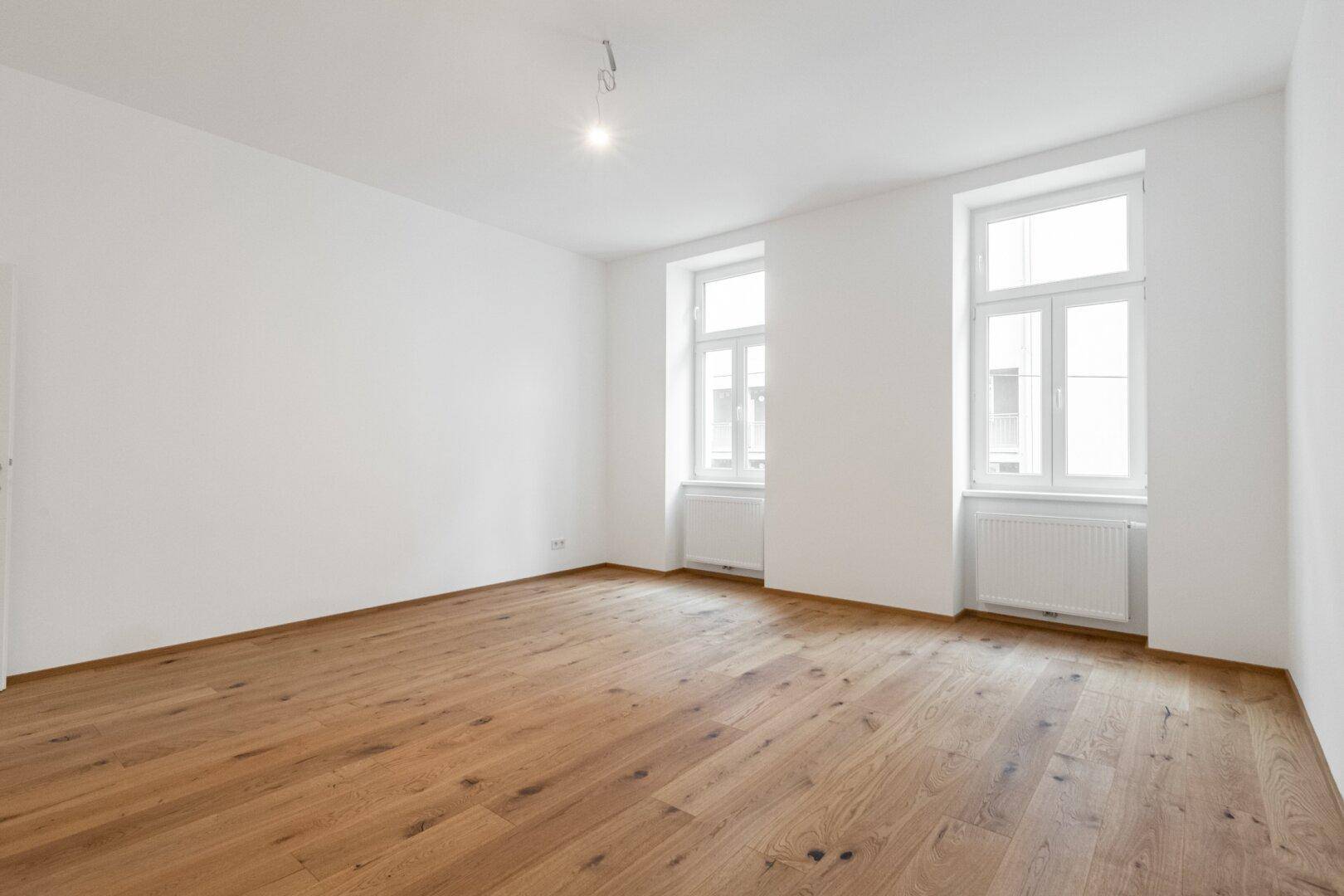 Leeres Wohnzimmer mit Holzboden und zwei Fenstern, bietet viel Potenzial für persönliche Einrichtungsideen.