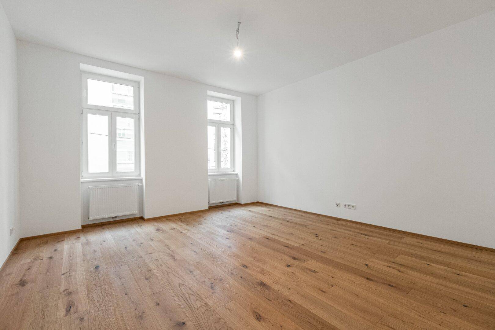 Geräumiges, unmöbliertes Wohnzimmer mit hellem Holzboden und zwei großen Fenstern, bereit zur individuellen Gestaltung.