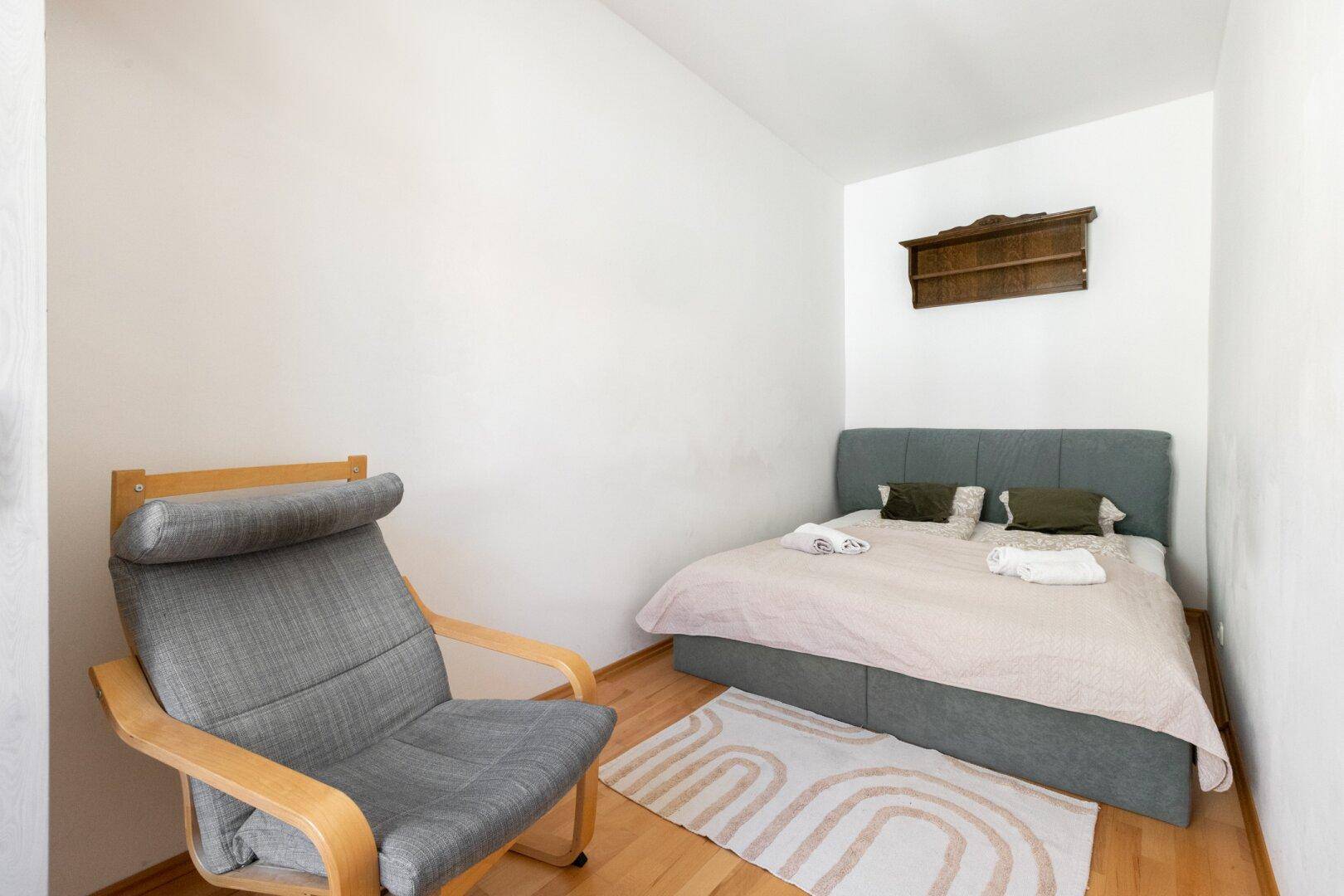 Kleines Schlafzimmer mit Einzelbett, Sessel und Holzboden in einem hellen Raum.
