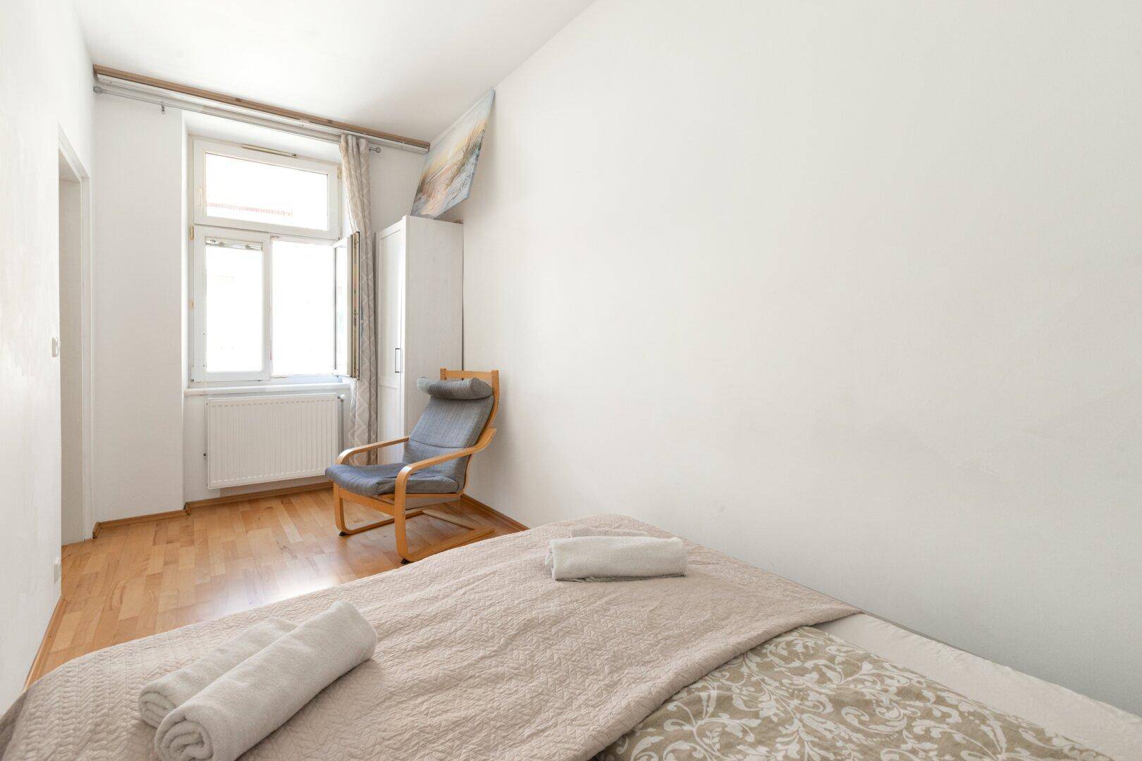 Helles Schlafzimmer mit Einzelbett, Sessel und Fenster, das viel Tageslicht hereinlässt.