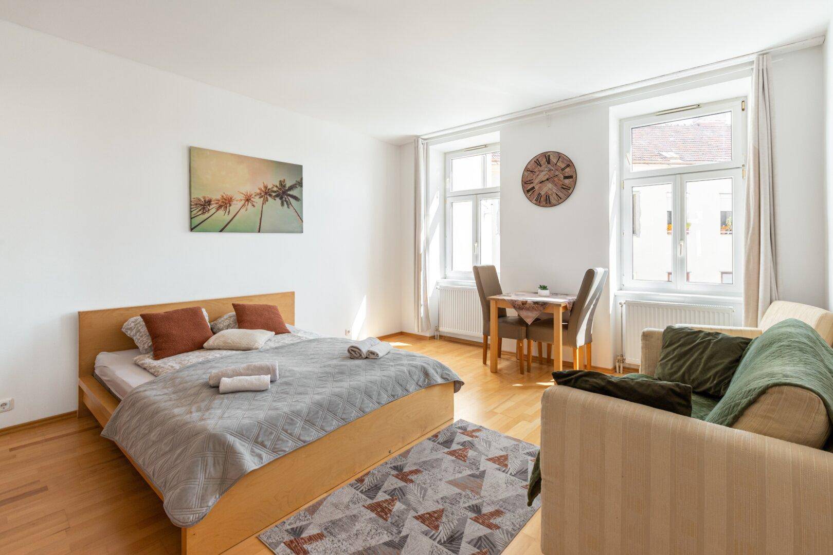 Schlafzimmer mit Holzboden, Doppelbett, Sofa und einem kleinen Esstisch am Fenster.