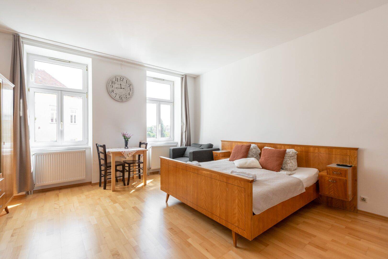 Helles Schlafzimmer mit Holzboden, klassischem Bett und einem kleinen Esstisch am Fenster.