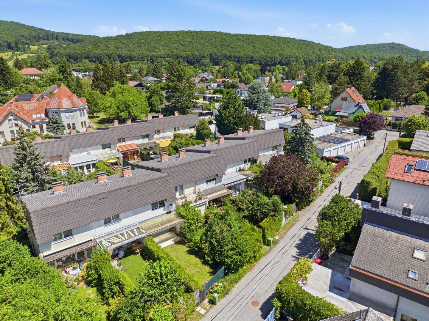 Weitläufige Luftaufnahme einer Wohnsiedlung mit Reihenhäusern und üppiger grüner Hügellandschaft im Hintergrund.