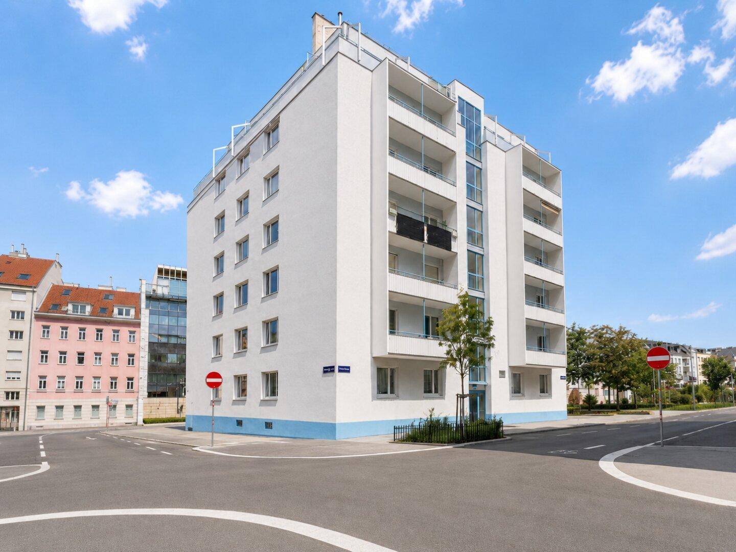 Modernes Wohngebäude mit weißer Fassade, blauen Akzenten und Balkonen an einer Straßenecke.