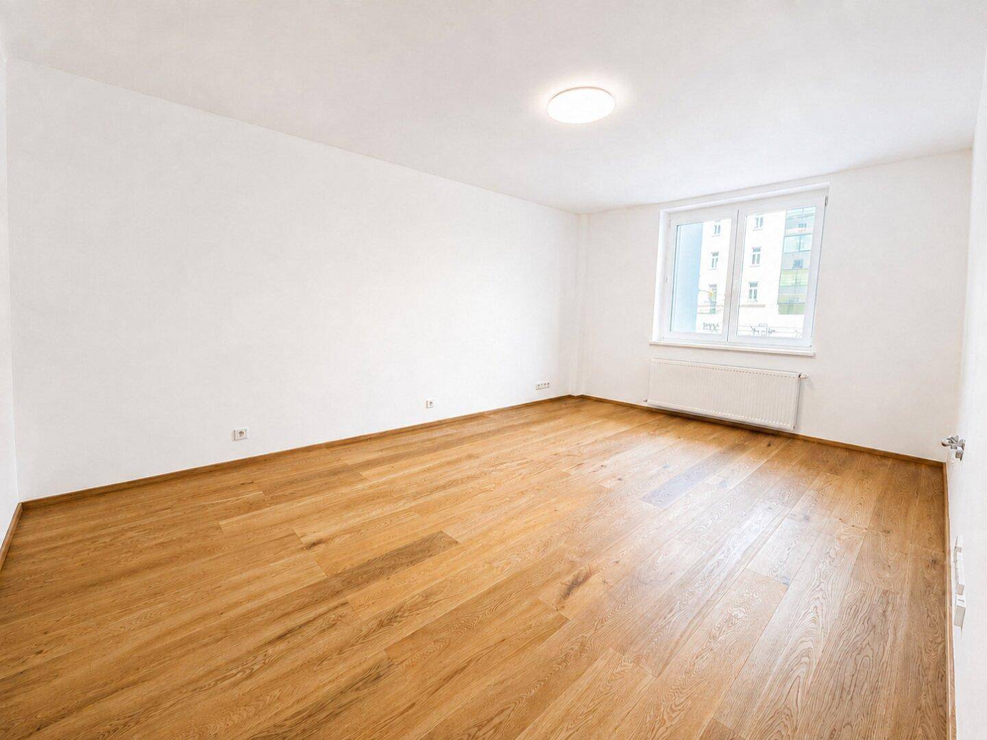 Geräumiger, unmöblierter Raum mit hellem Holzparkett und einem Fenster.