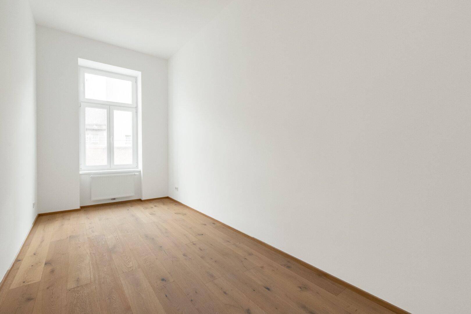 Leeres Schlafzimmer mit hellem Parkettboden und einem Fenster.