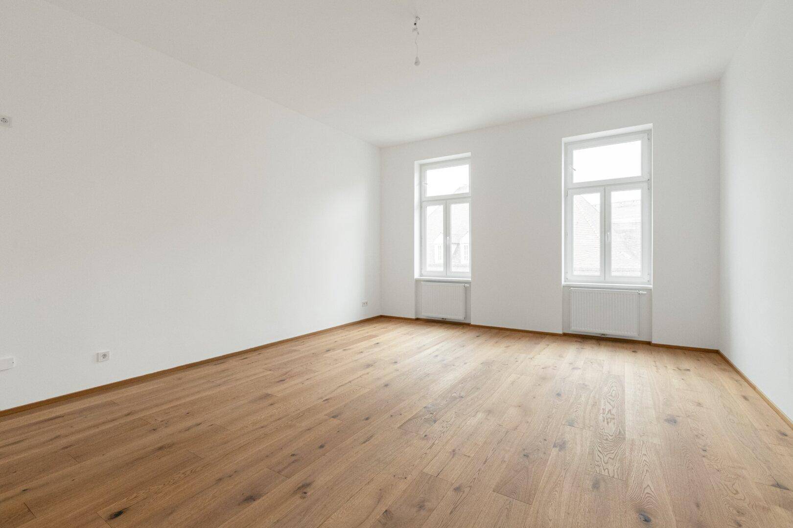 Heller, leerer Wohnraum mit zwei großen Fenstern und Holzboden.