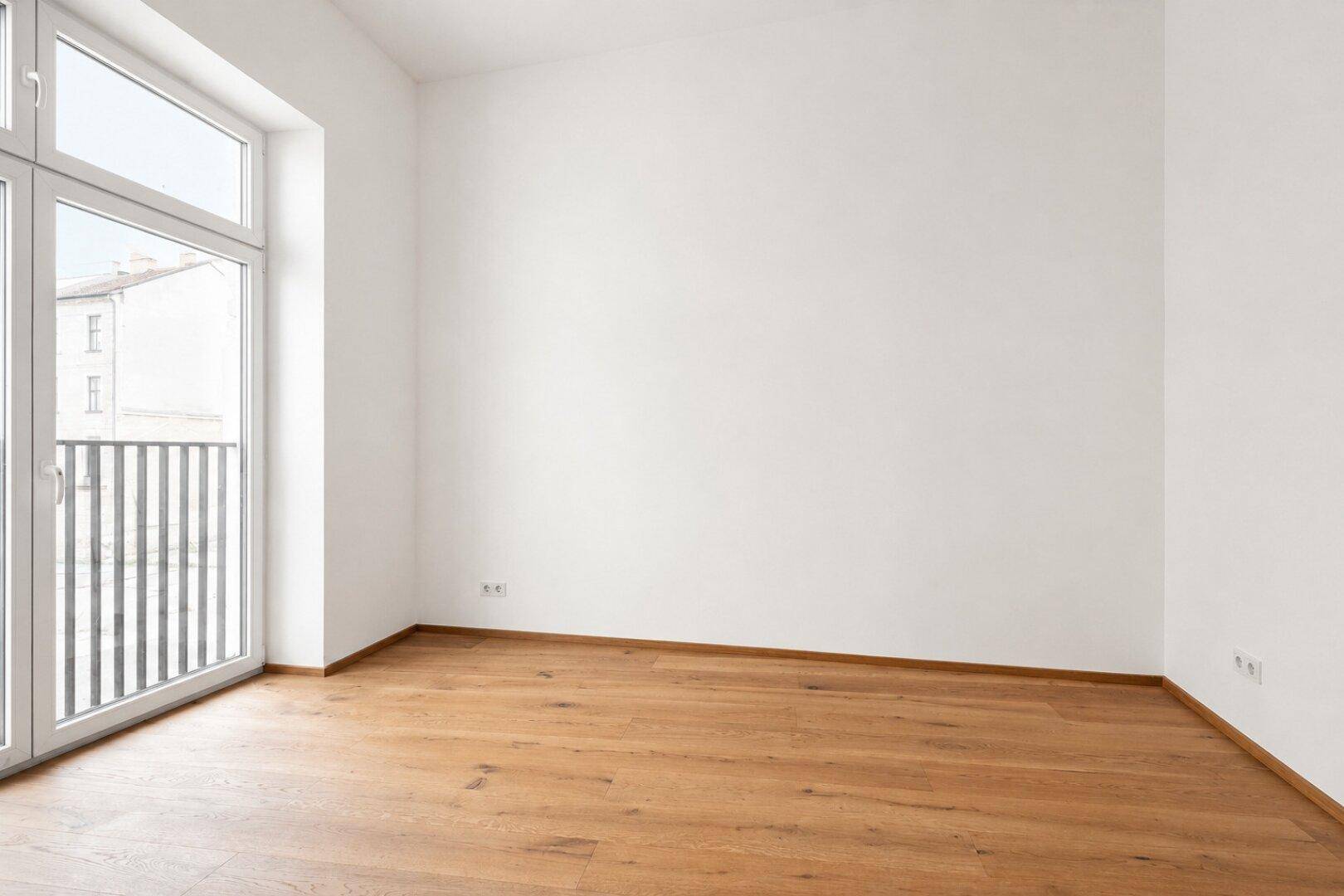 Geräumiges, leeres Zimmer mit Holzboden und großem Fenster zum französischen Balkon.