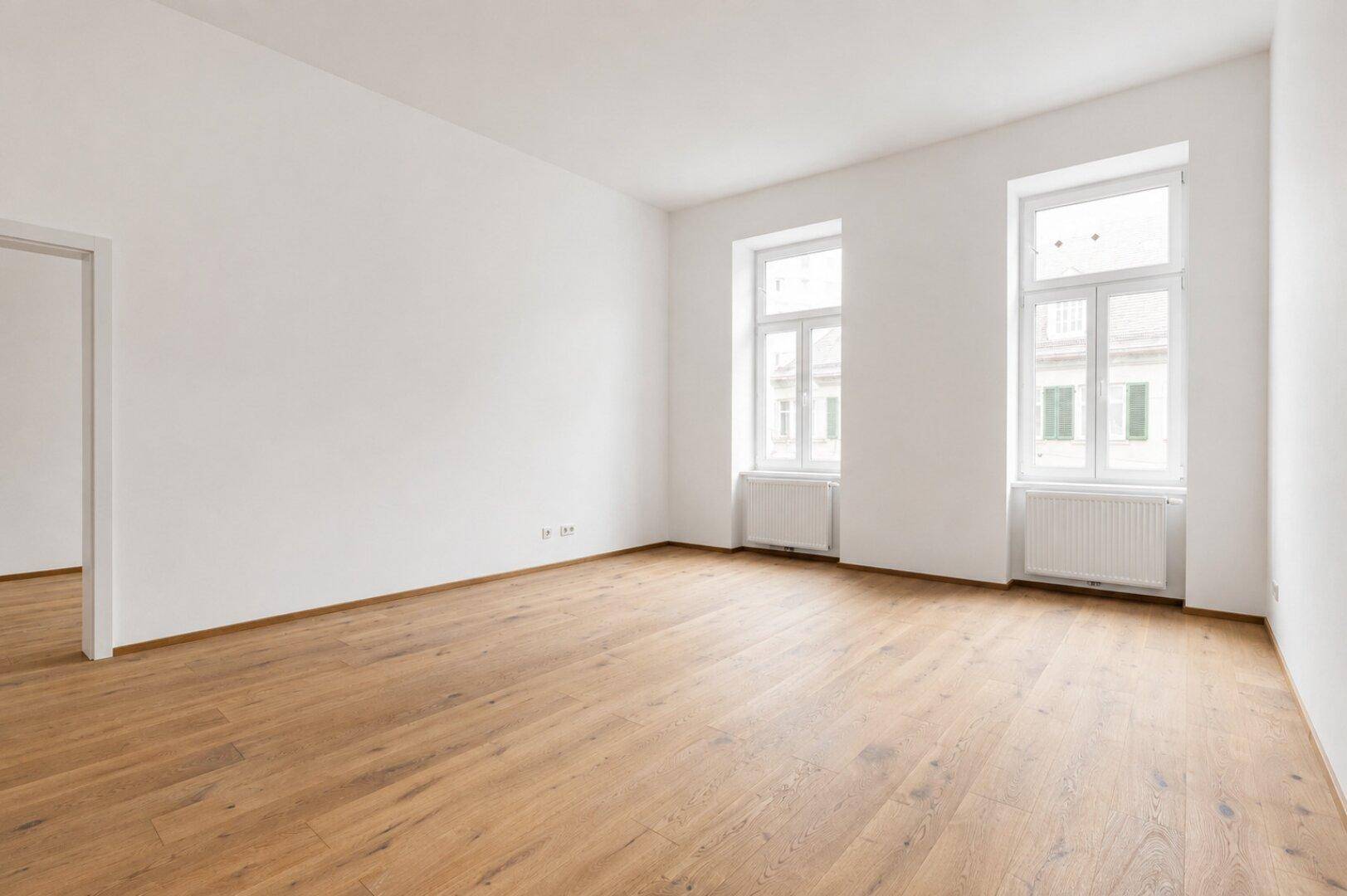 Geräumiges, leeres Wohnzimmer mit hellem Holzboden und zwei großen Fenstern.