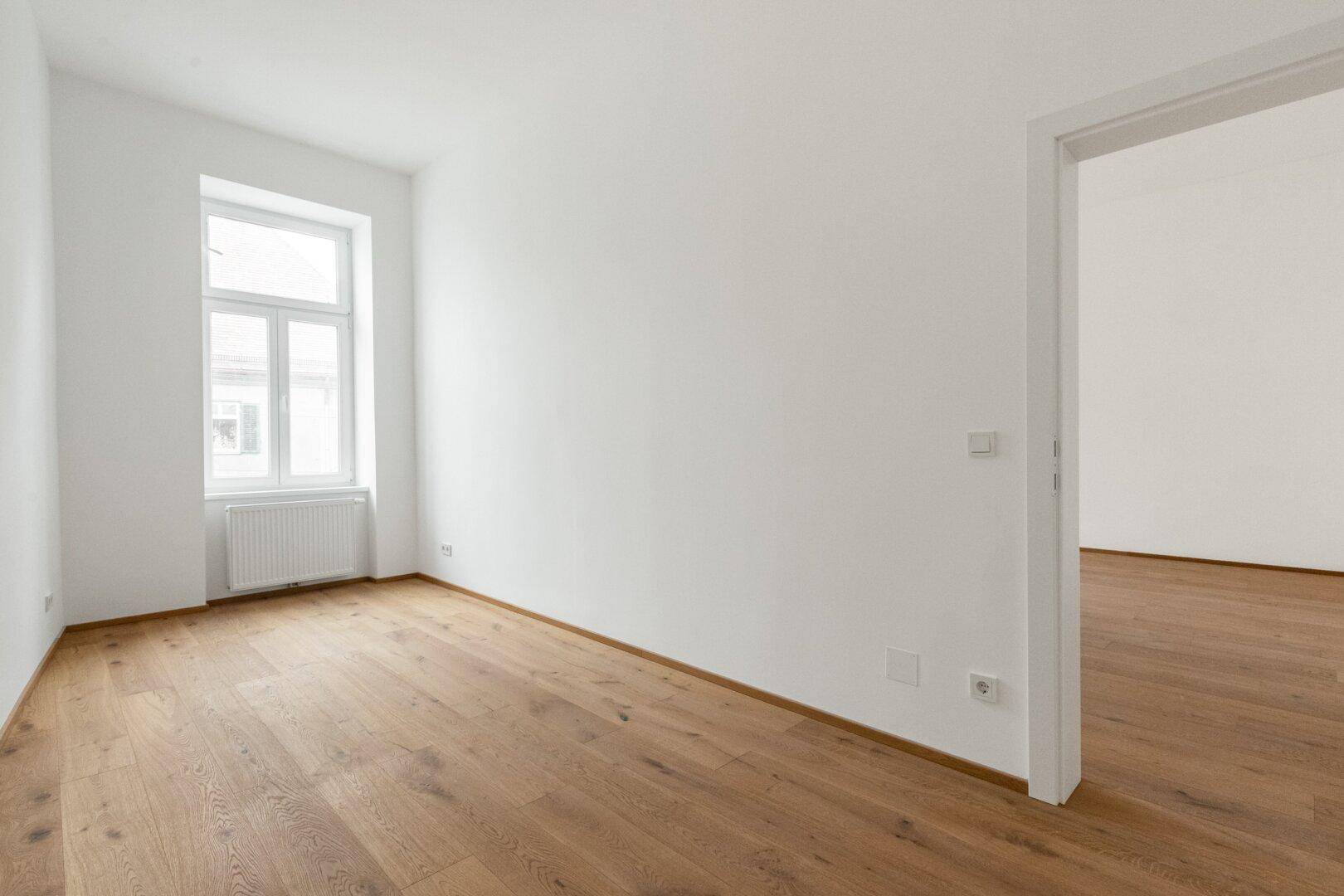 Ruhiges, leeres Zimmer mit Holzboden und einem Fenster, ideal als Schlaf- oder Arbeitszimmer.