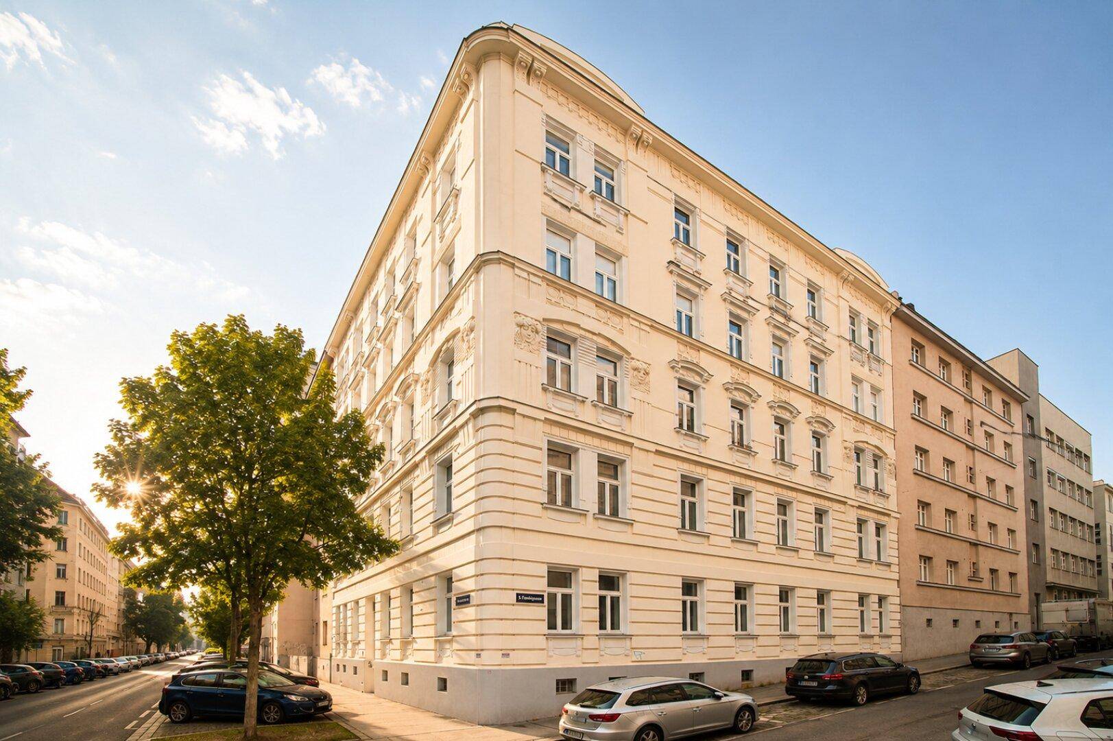 Historisches Eckgebäude mit klassischer Architektur und vielen Fenstern in einer sonnigen städtischen Umgebung.