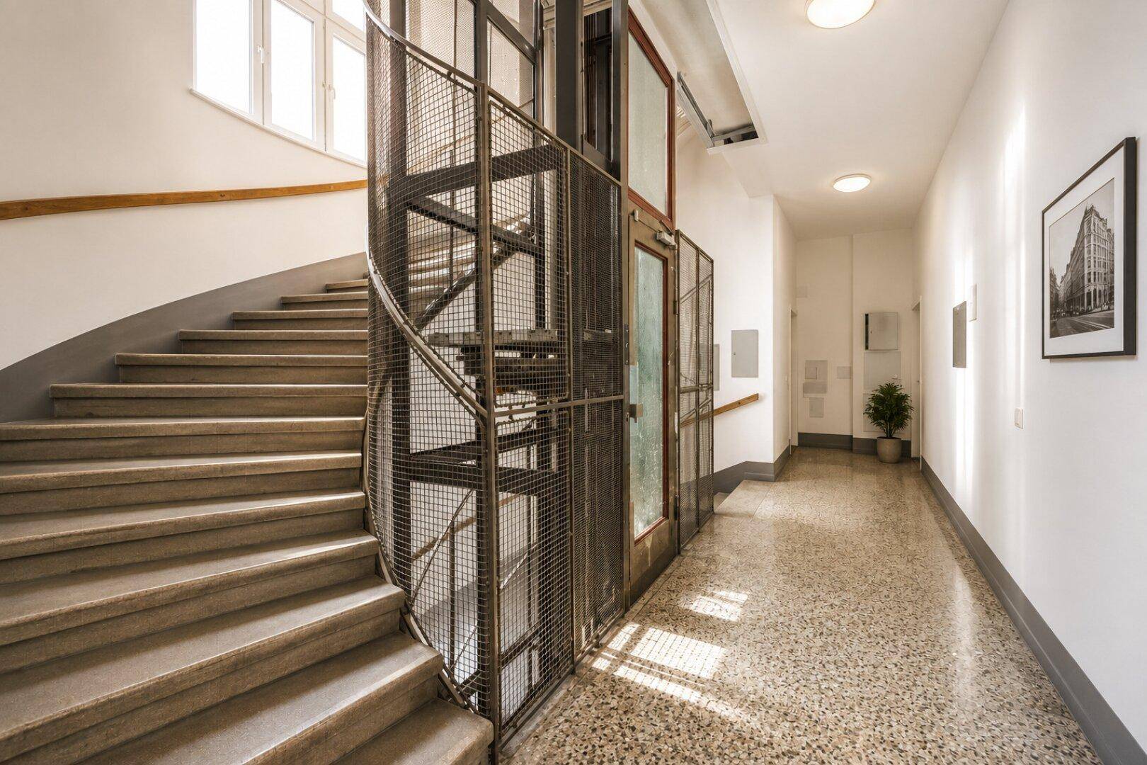 Heller Flur mit Steintreppe und einem modernen Gitteraufzug in einem Altbau.