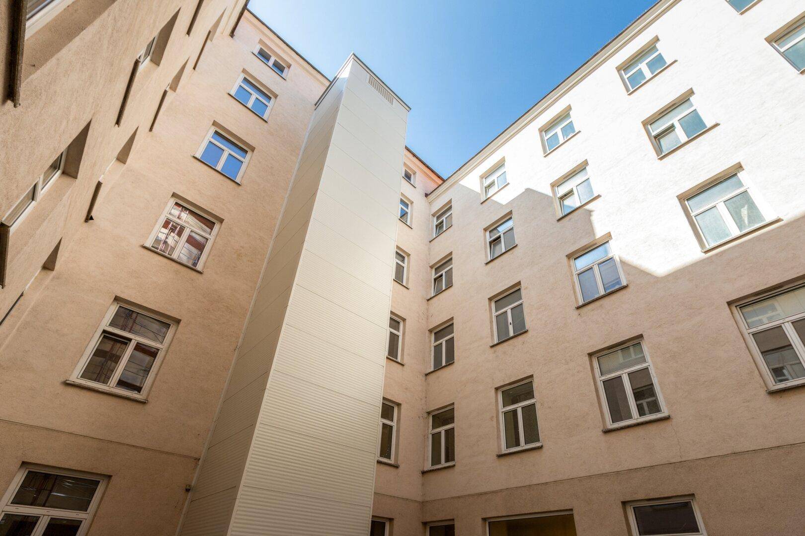 Blick in den hellen Innenhof eines Wohngebäudes mit vielen Fenstern und einem Aufzugsschacht.