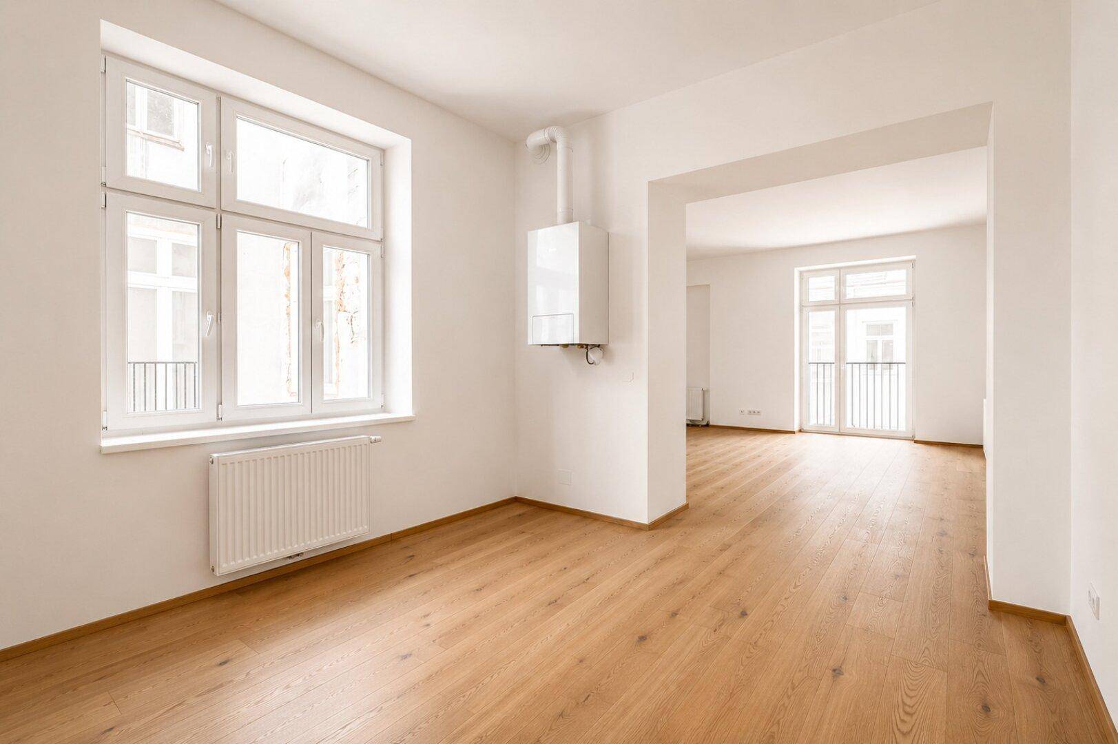Unmöblierter Wohnbereich mit Holzboden, großen Fenstern und Heizungsanlage an der Wand.