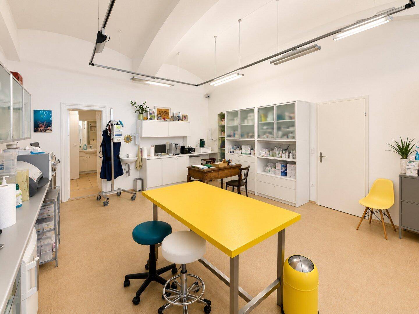 Heller Behandlungsraum mit gelbem Tisch, medizinischer Ausstattung und gewölbter Decke.