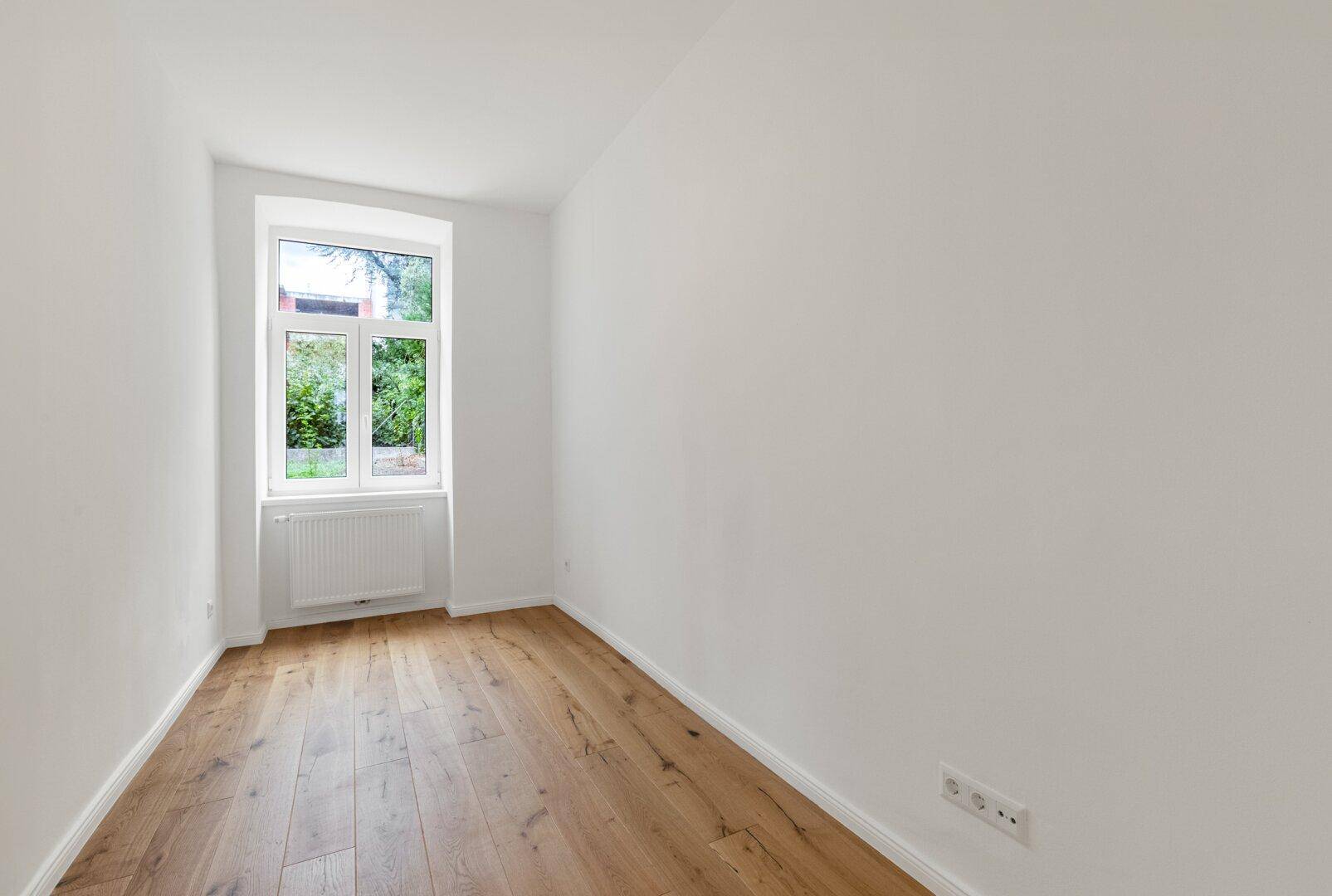 Kleines, helles Zimmer mit Holzboden und einem Fenster, ideal als Schlaf- oder Arbeitszimmer.