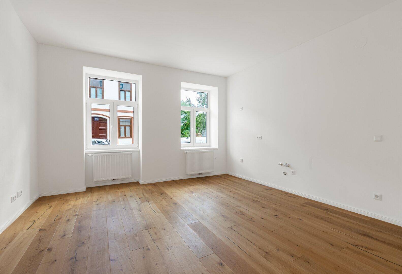 Helles, leeres Zimmer mit Holzboden und zwei Fenstern, bereit zur individuellen Gestaltung.