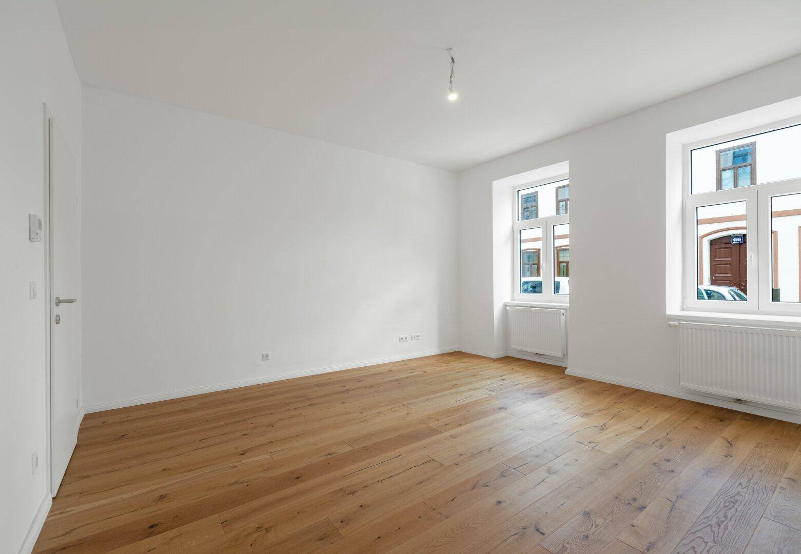Leeres, helles Zimmer mit Holzboden und zwei Fenstern, ideal für Wohn- oder Schlafbereich.