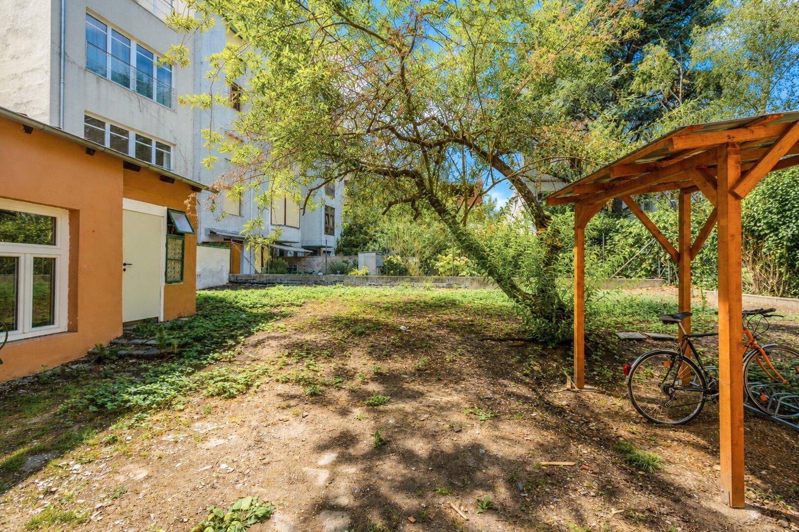 Gartenbereich mit Fahrradunterstand und altem Baum, umgeben von Gebäuden und Grün.