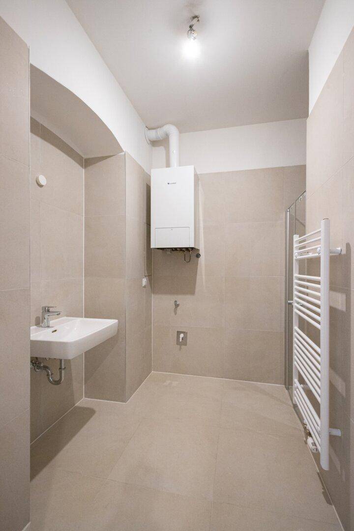 Modernes Badezimmer mit beigefarbenen Fliesen, Waschbecken, Heiztherme und Handtuchheizkörper.