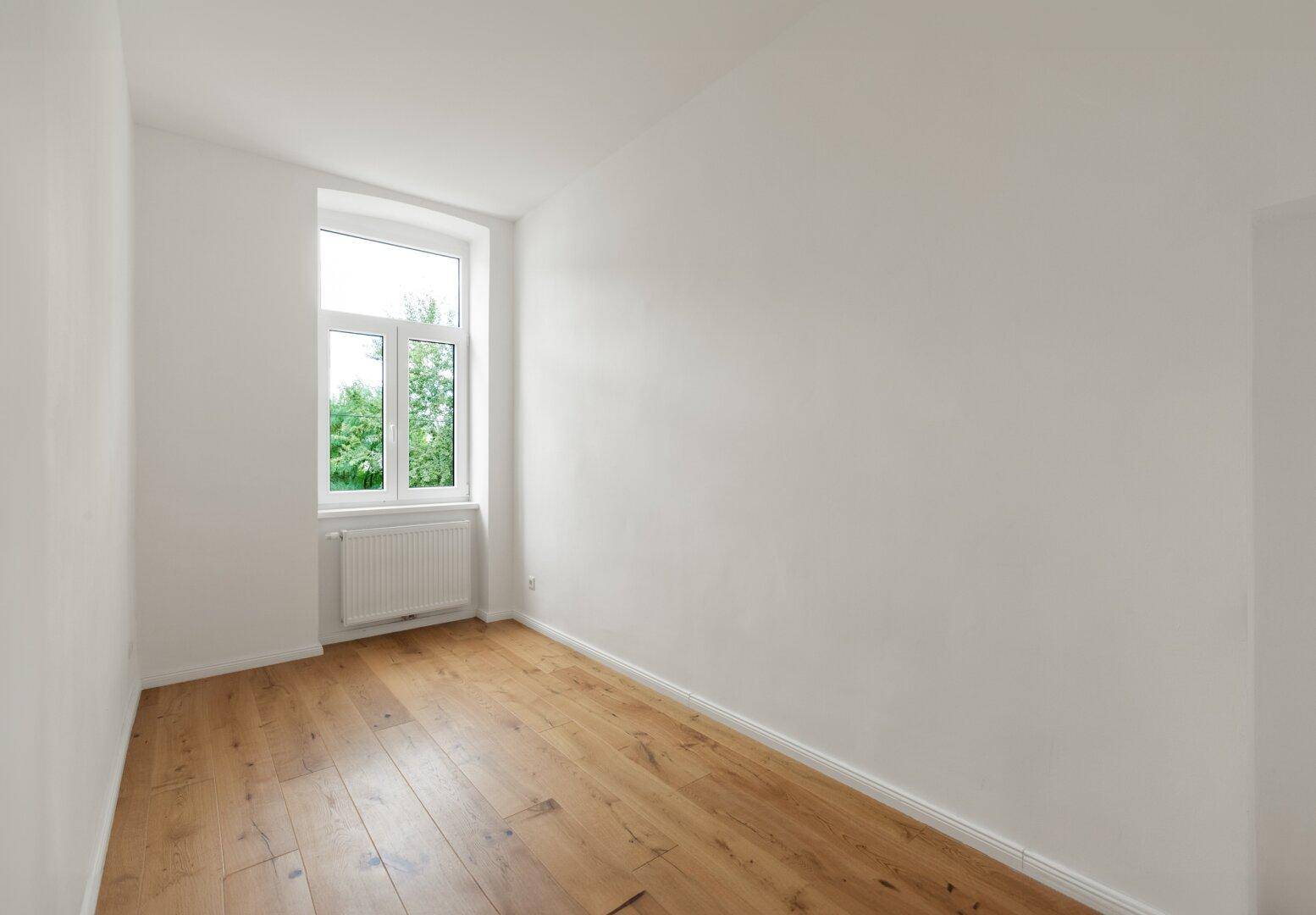 Kleineres, leeres Zimmer mit Holzboden, weißen Wänden und einem Fenster mit Grünblick.