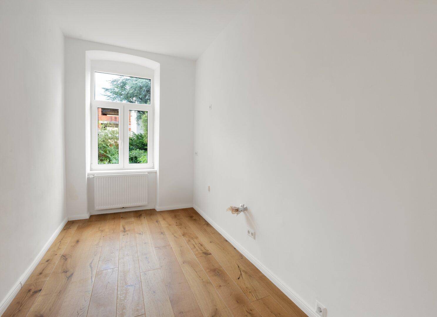 Leerer Küchenraum mit Holzboden, hellem Fenster und vorbereiteten Anschlüssen für die Kücheninstallation.