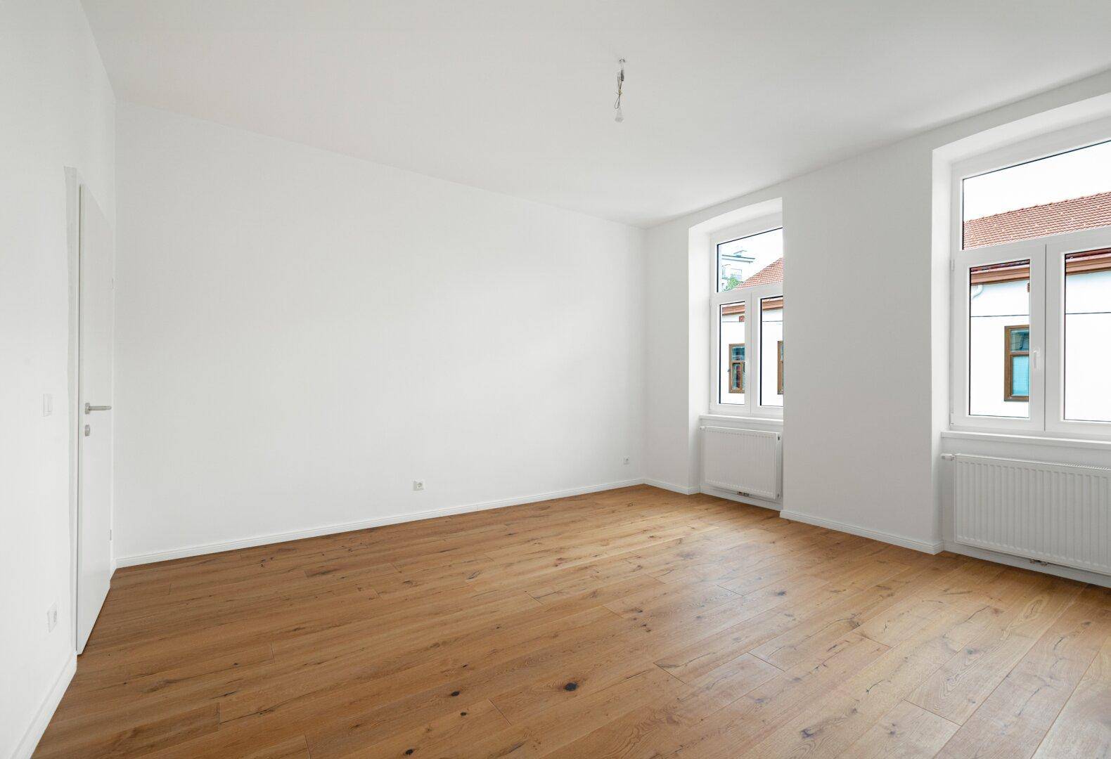 Helles, leeres Zimmer mit Holzboden und zwei Fenstern, ideal als Schlaf- oder Arbeitszimmer nutzbar.