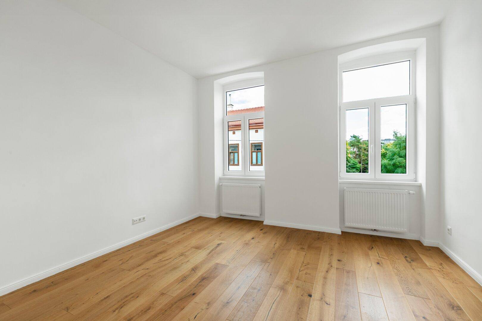 Geräumiges, leeres Zimmer mit Holzboden, weißen Wänden und zwei großen Fenstern, vielseitig nutzbar.