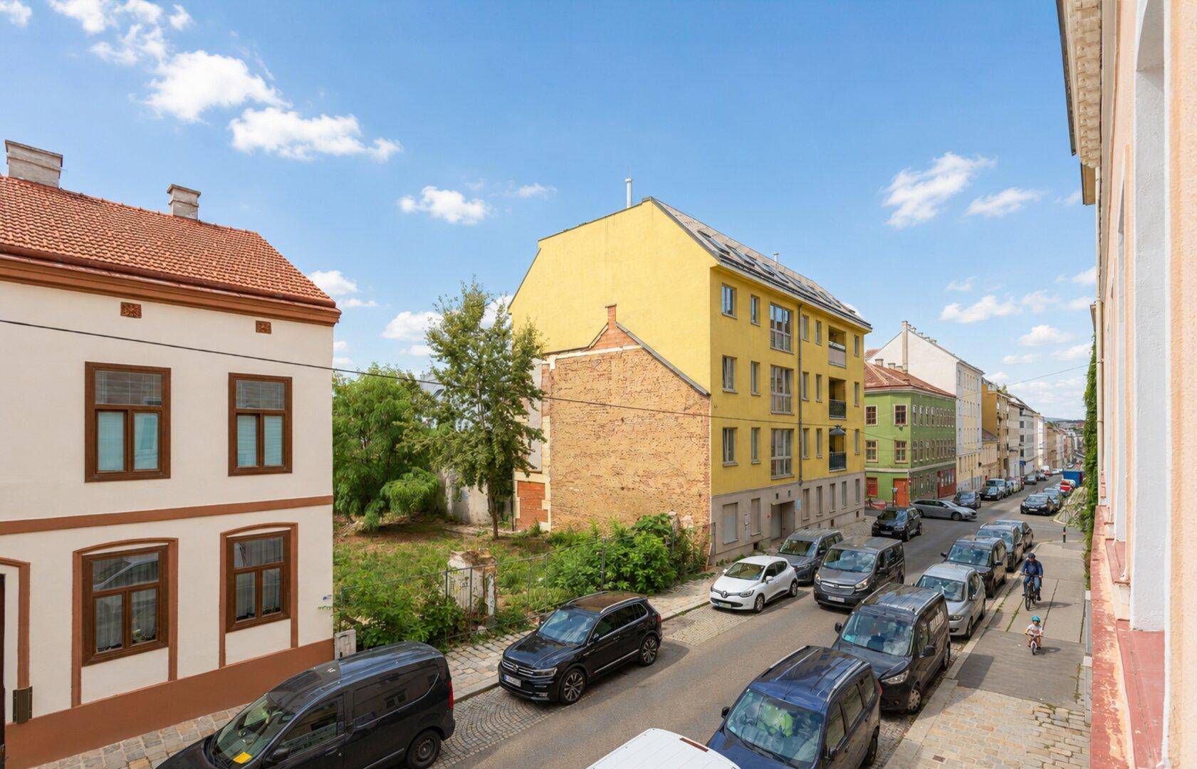 Blick auf eine belebte Straße mit parkenden Autos und angrenzenden Gebäuden unter blauem Himmel.