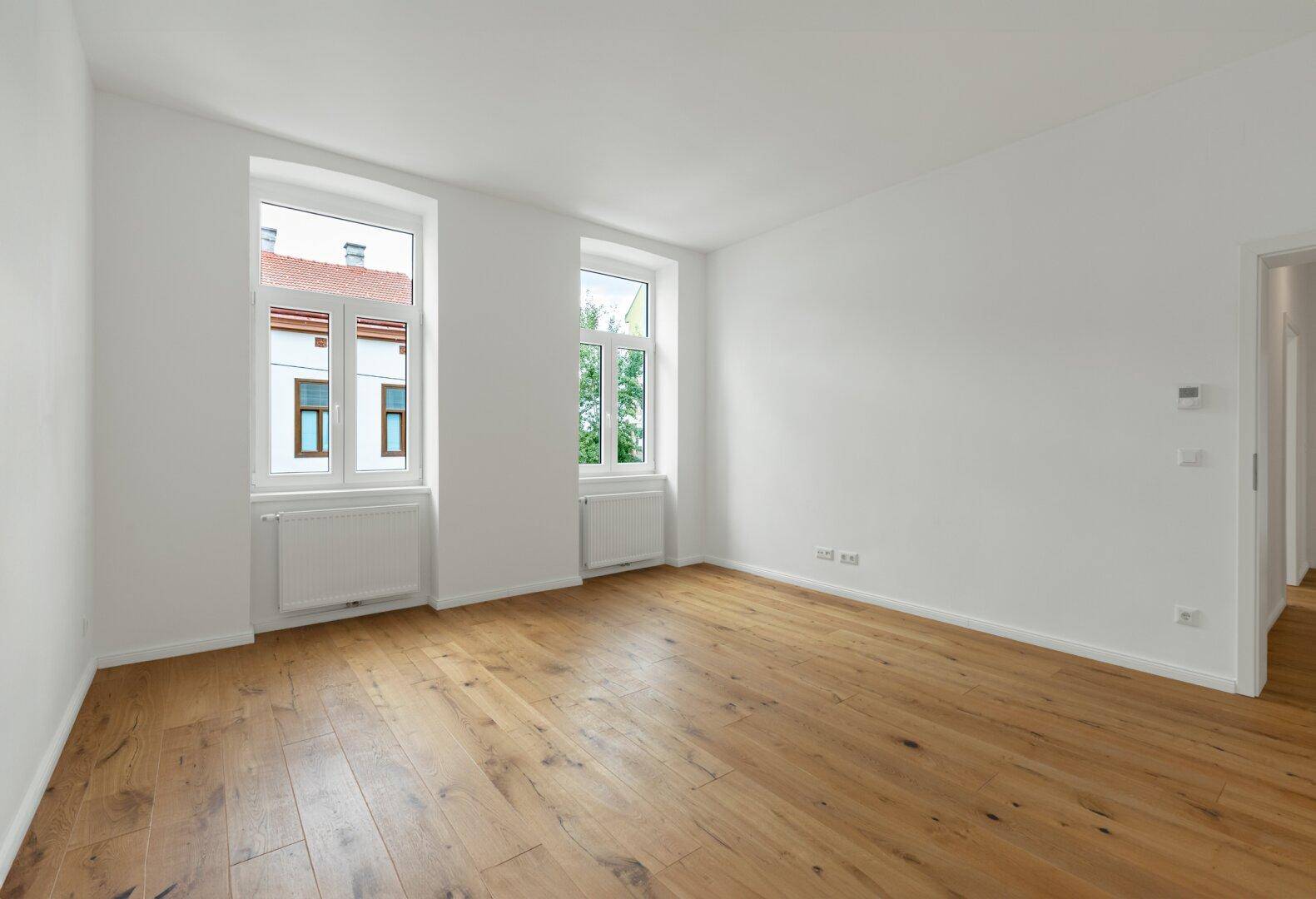 Geräumiges, leeres Zimmer mit hellen Wänden, Holzboden und zwei großen Fenstern, bereit zur Gestaltung.
