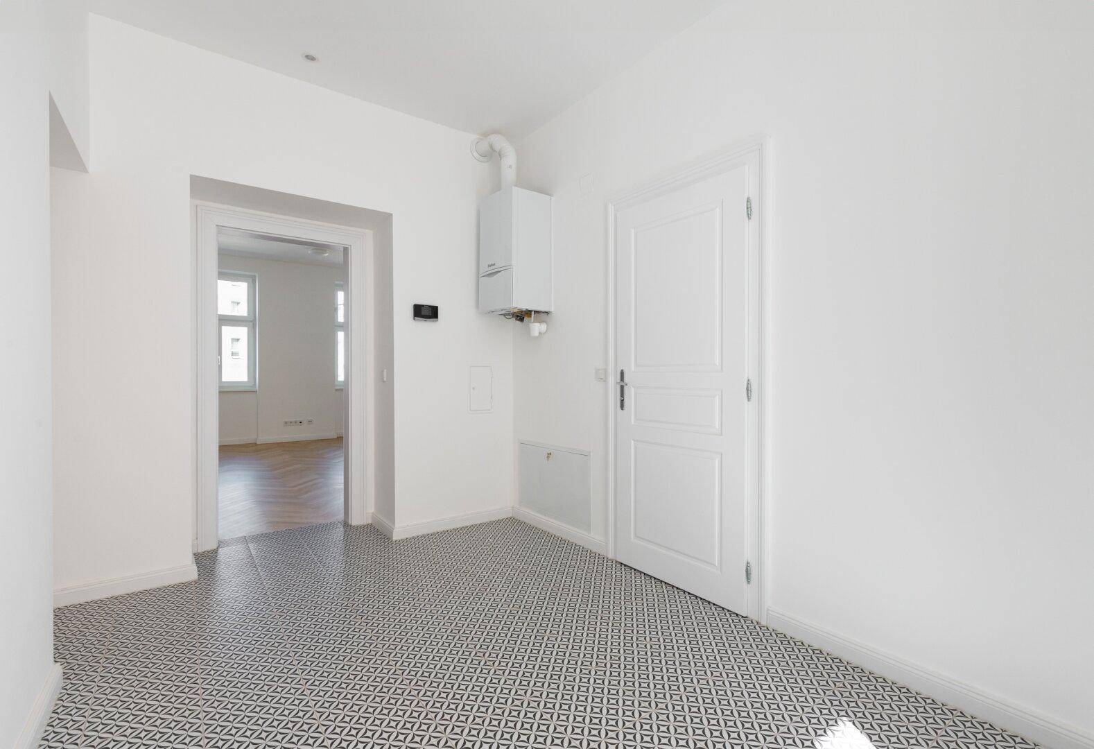 Flur mit gemusterten Fliesen, weißer Tür und sichtbarem Heizkessel an der Wand.