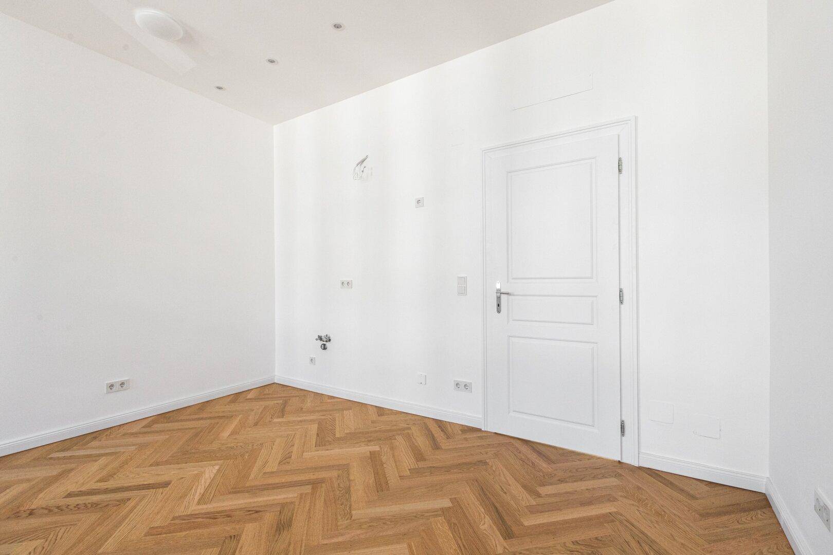Leeres Zimmer mit Fischgrätparkett, weiß gestrichenen Wänden und einer schlichten Tür.
