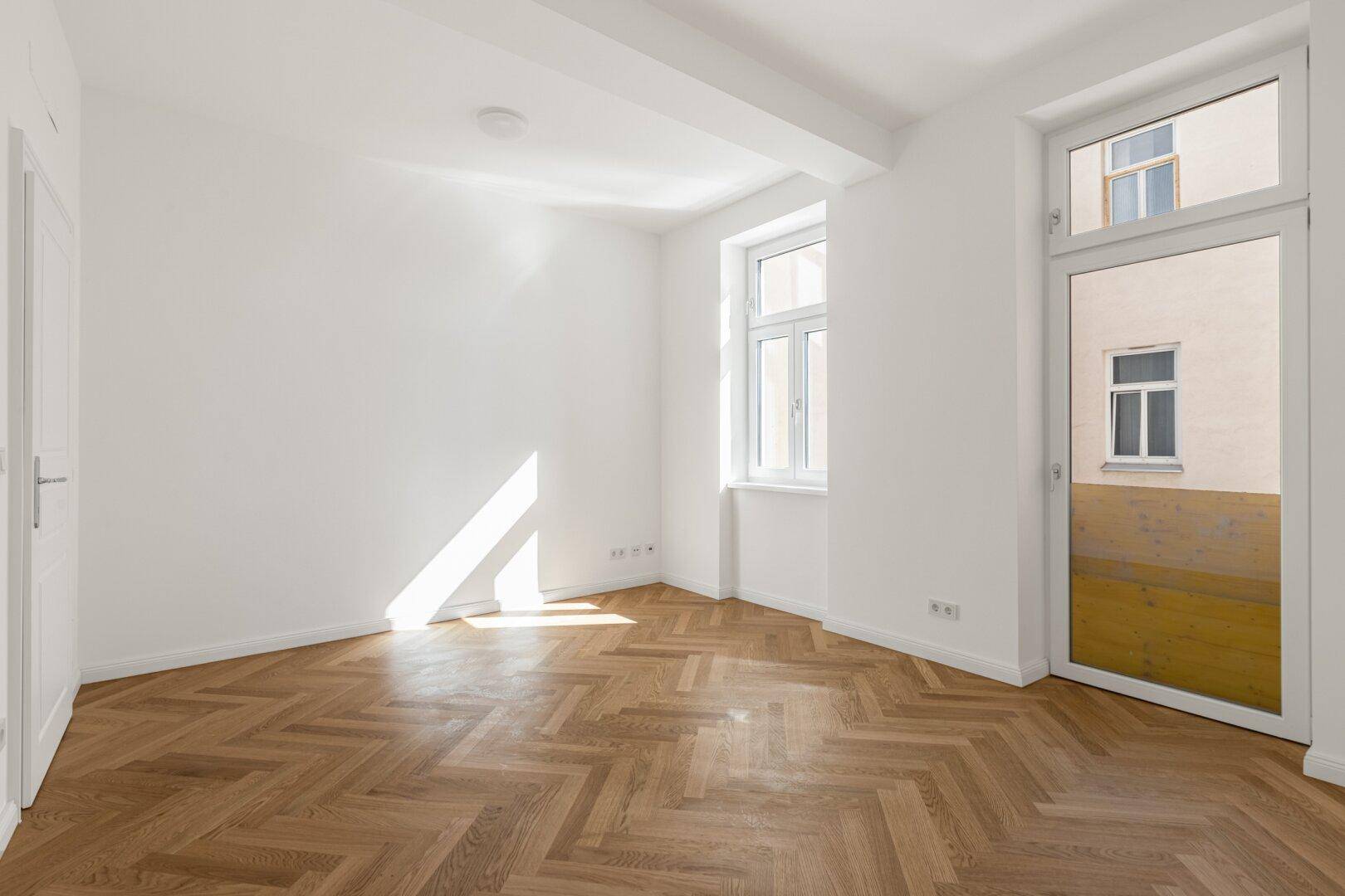 Lichtdurchflutetes Zimmer mit Fischgrätparkett und Zugang zu einem potenziellen Balkon oder Terrasse.