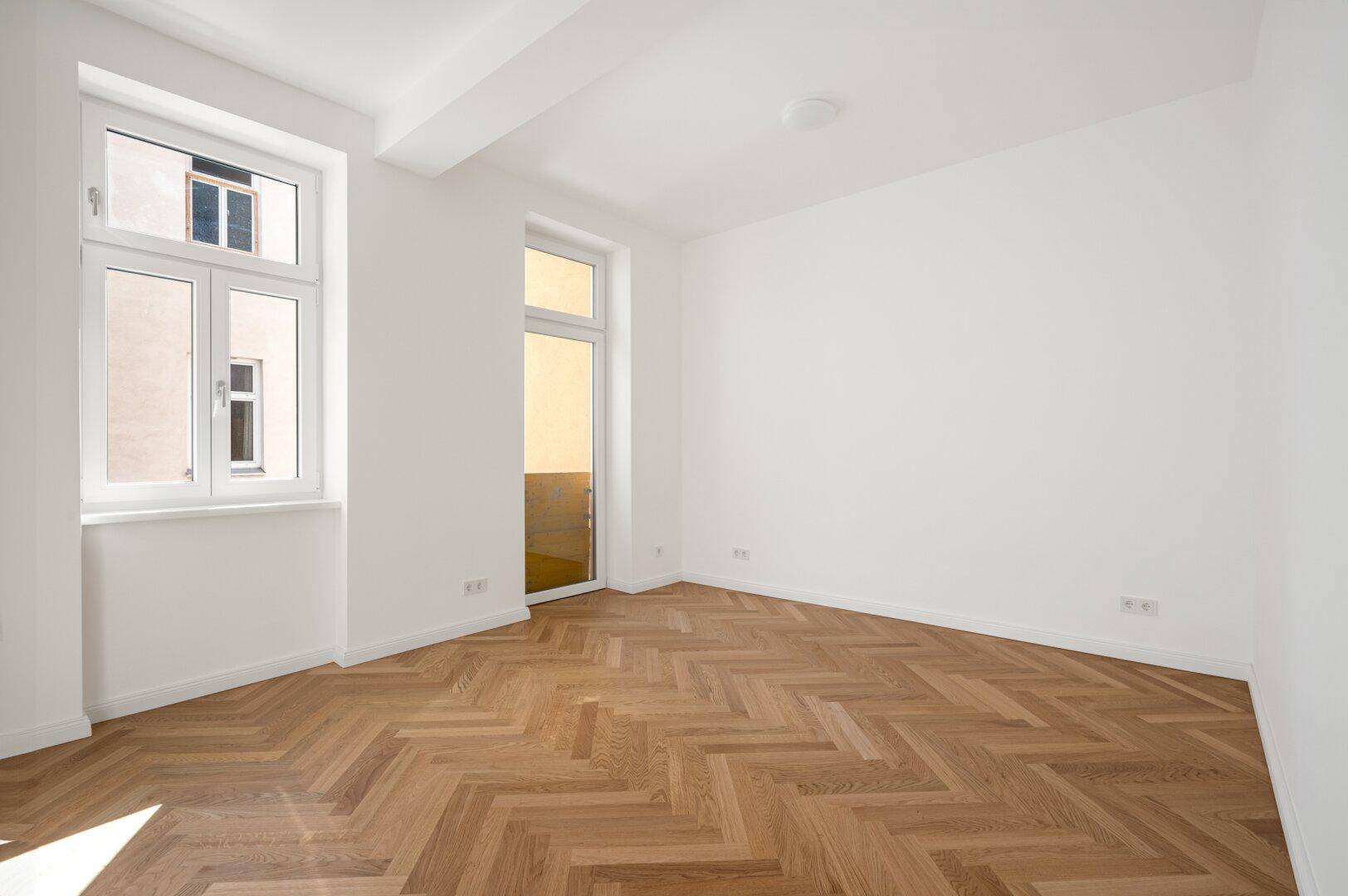 Geräumiges Zimmer mit Fischgrätparkett, großen Fenstern und viel natürlichem Licht.