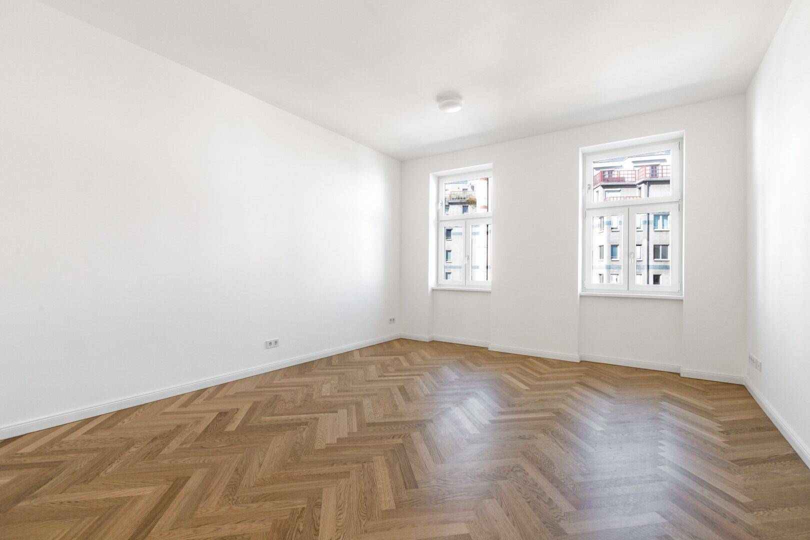 Geräumiges Zimmer mit Fischgrätparkett und zwei Fenstern, die viel Tageslicht hereinlassen.
