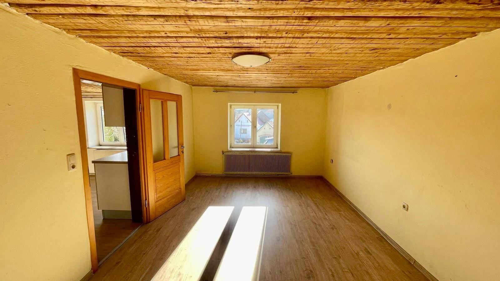 Leeres Wohnzimmer mit Holzbalkendecke, Laminatboden und Fenster mit Tageslicht.
