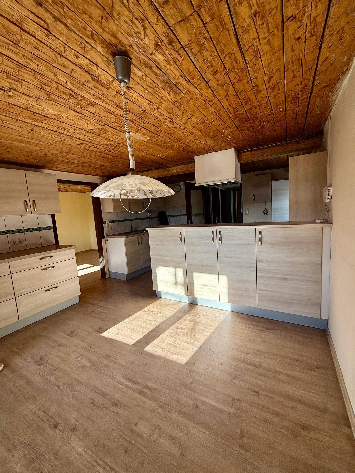 Helle Küche mit hellen Holzschränken, Holzbalkendecke und Laminatboden, durchflutet von Sonnenlicht.