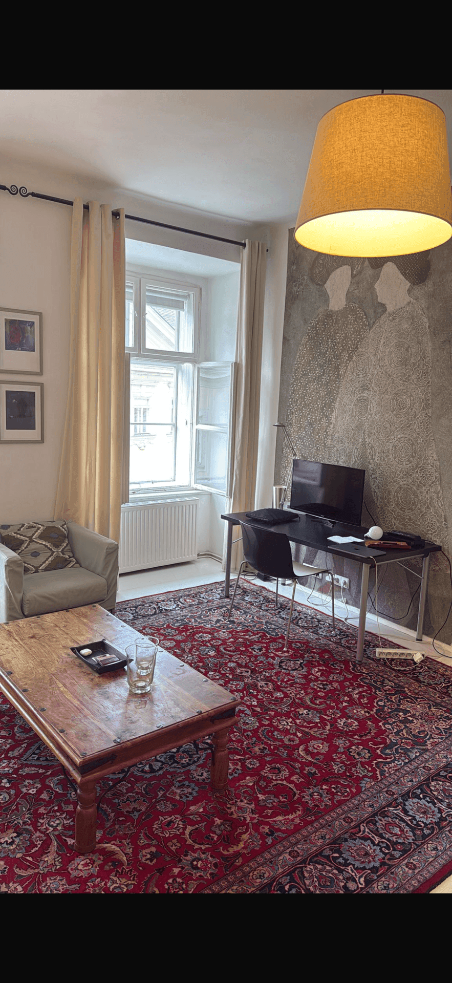 Heller Wohnbereich mit Schreibtisch, großem Fenster und einem auffälligen roten Orientteppich.
