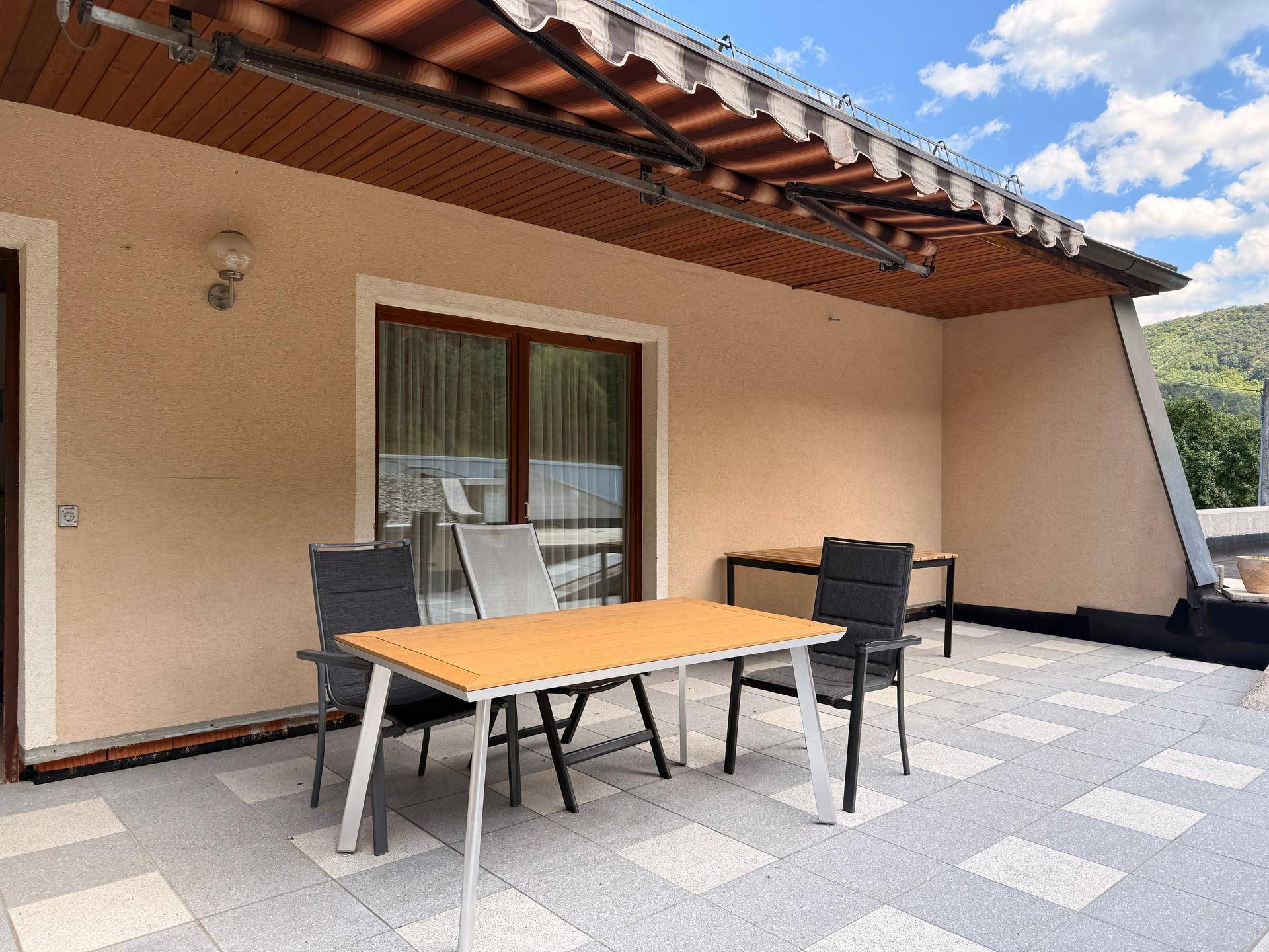 Geräumige Terrasse mit Fliesenboden, Markise und Sitzgelegenheiten, umgeben von grüner Landschaft.