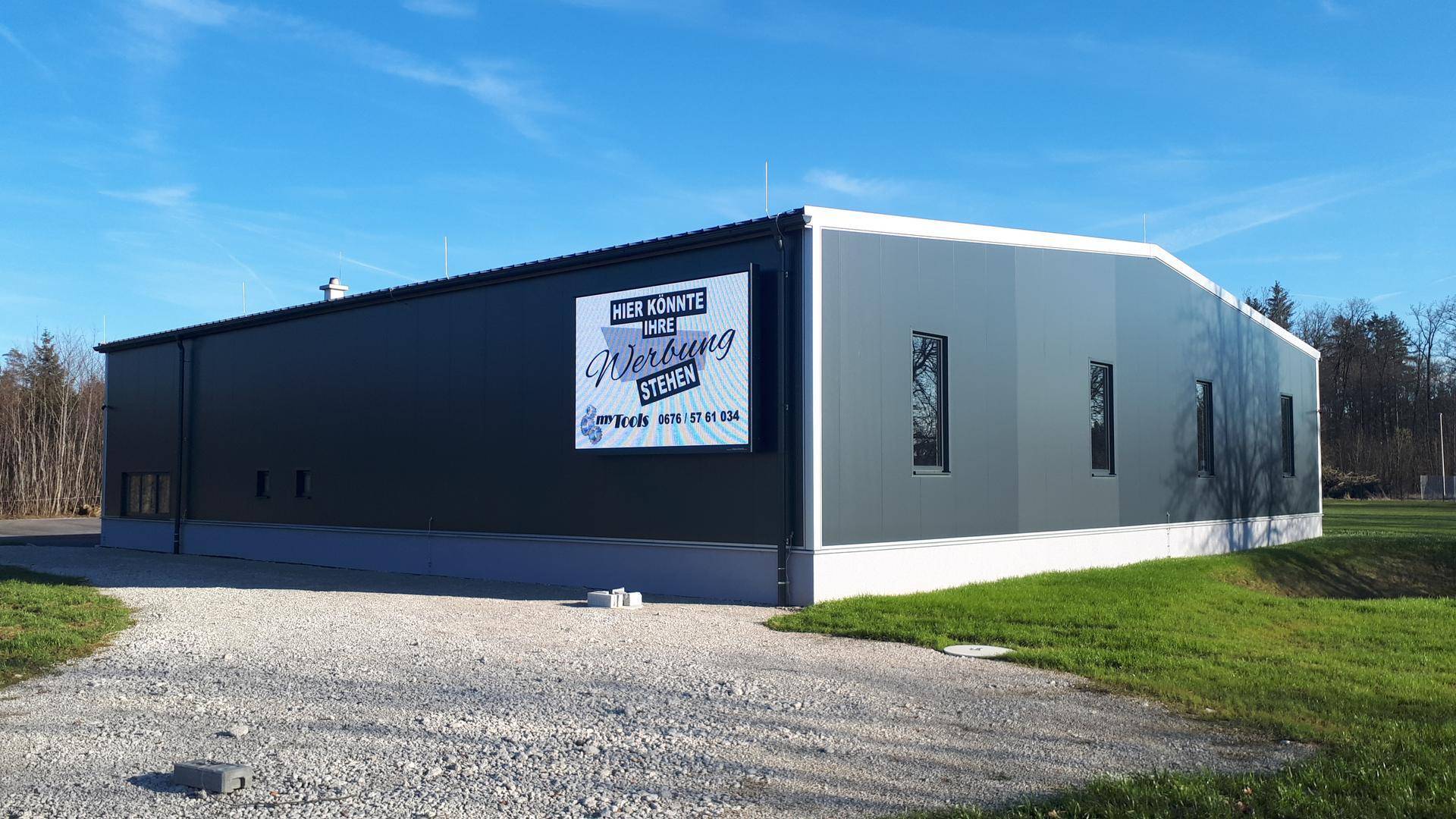 Modernes Gewerbegebäude mit dunkler Fassade und Werbefläche, umgeben von Grünflächen unter blauem Himmel.