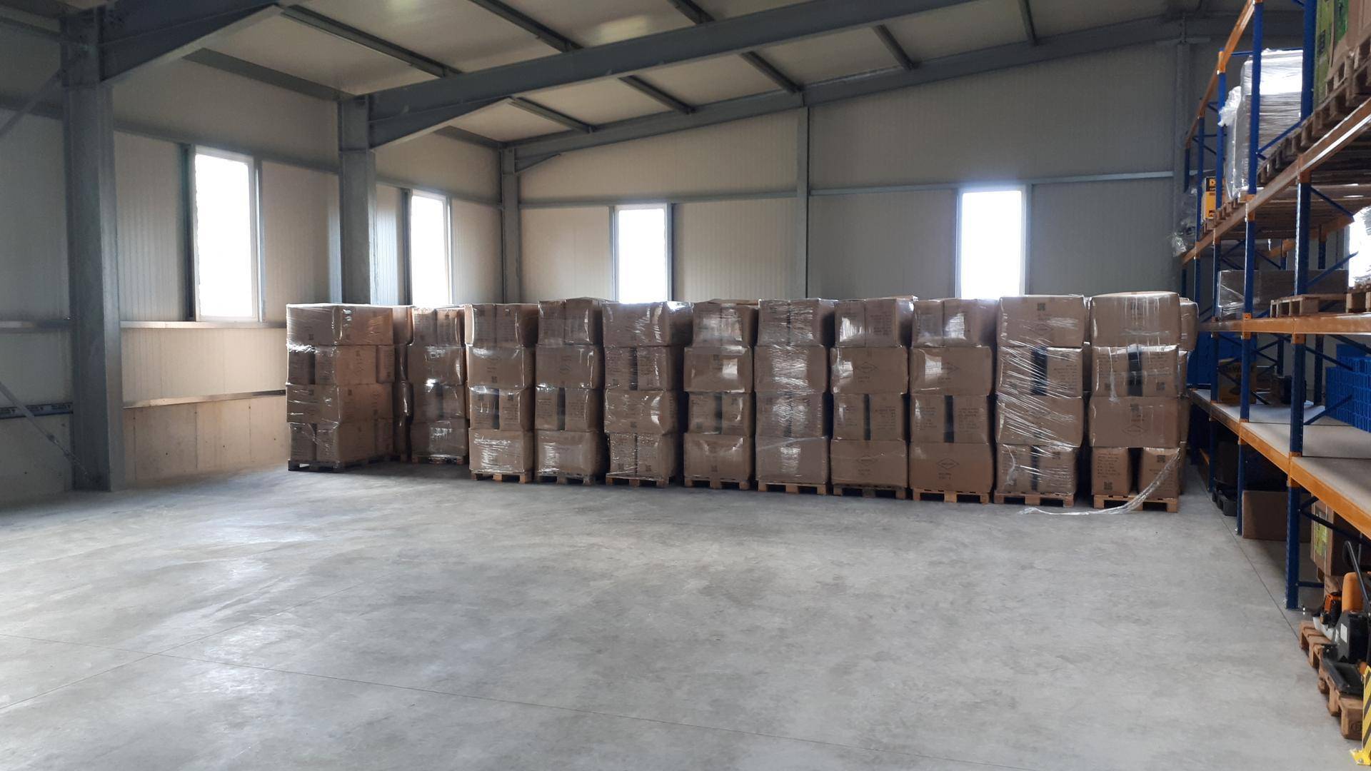 Großer Lagerraum mit gestapelten Kartons, hohen Decken und mehreren Fenstern, die Tageslicht hereinlassen.