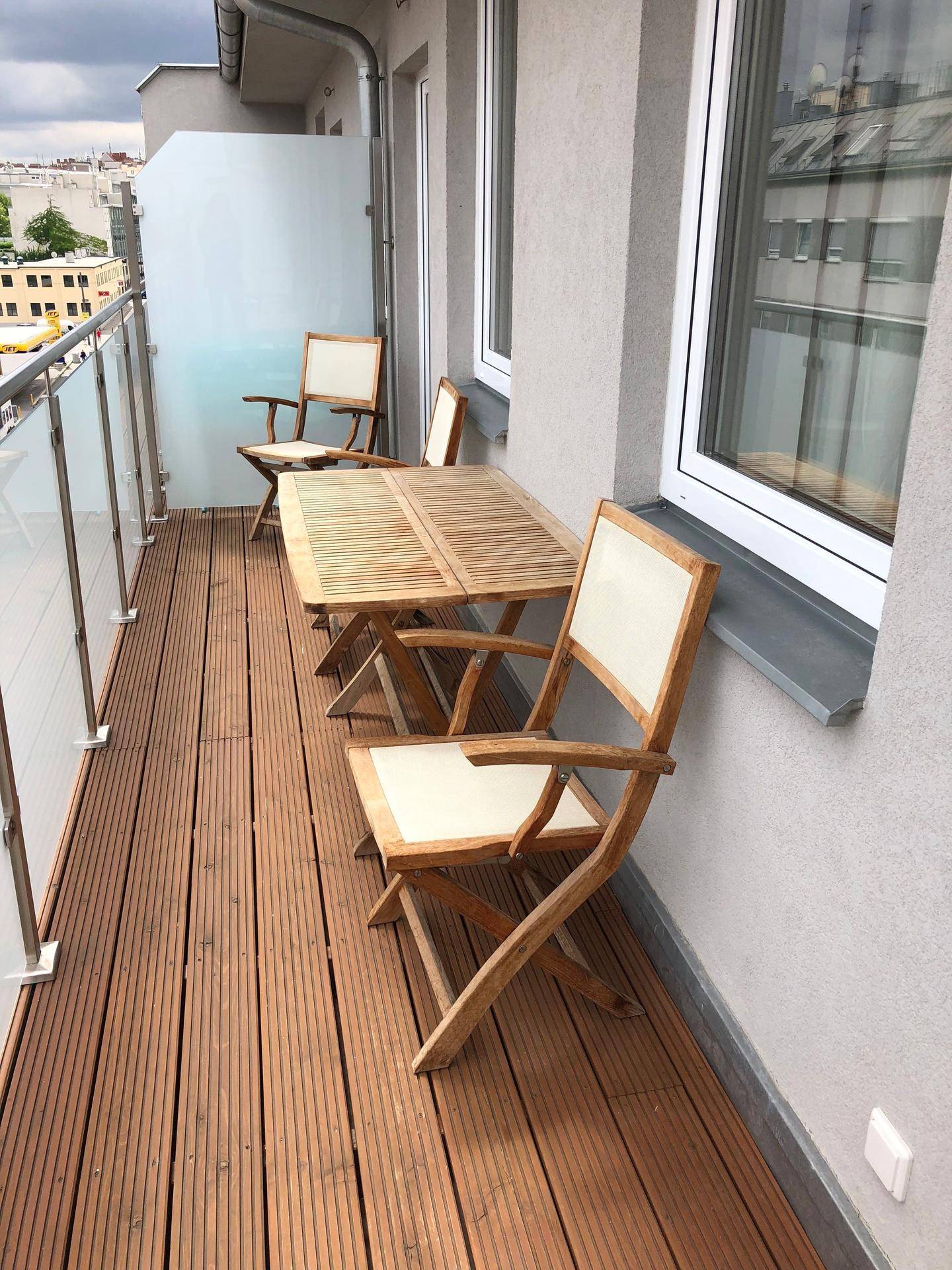 Schmaler Balkon mit Holzboden, Glasgeländer und Gartenmöbeln.