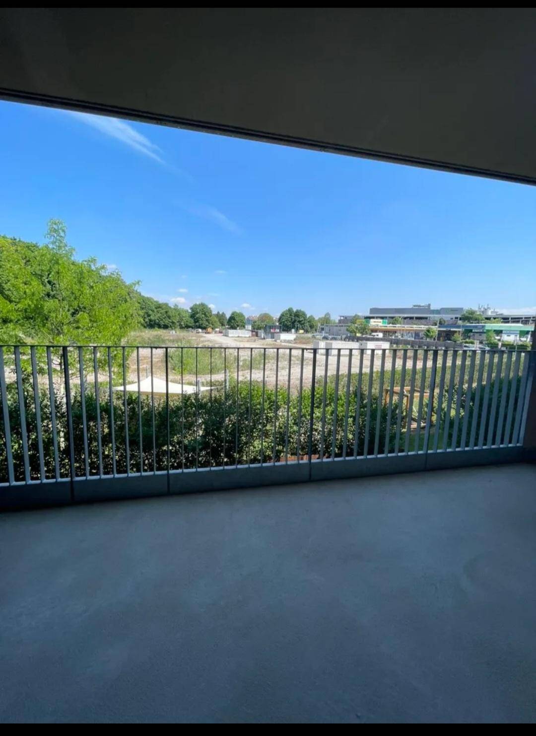 Balkon mit Blick auf eine Grünfläche und umliegende Gebäude unter blauem Himmel.
