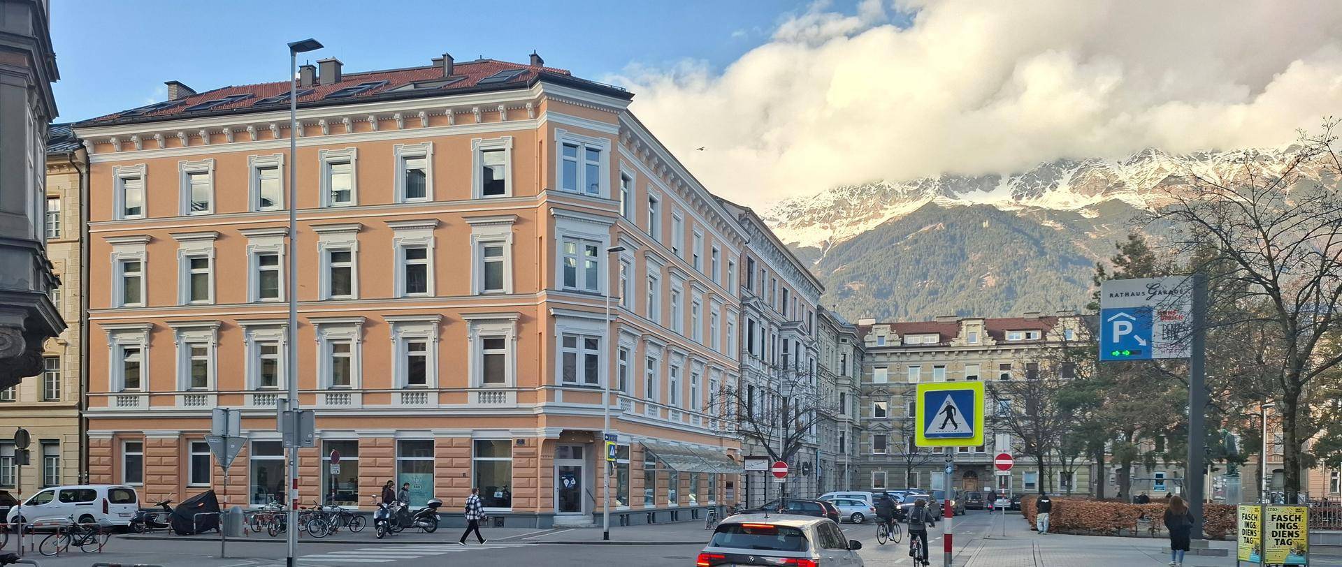 Historisches Gebäude mit klassischer Fassade an einer belebten Straße und schneebedeckten Bergen im Hintergrund.
